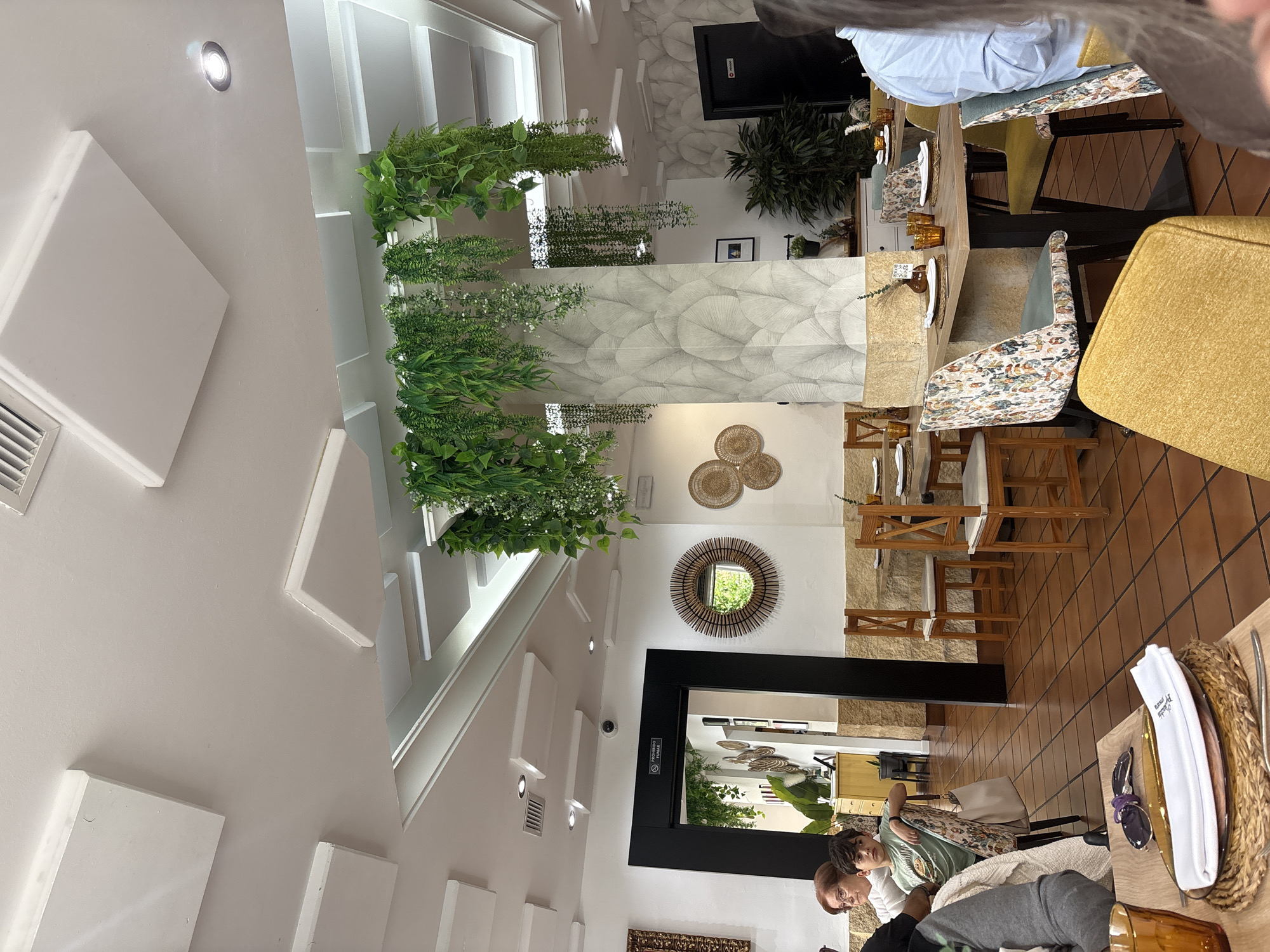 The image shows the interior of a restaurant in Córdoba, Spain. The restaurant features a modern decor with white walls accented by geometric white blocks and hanging plants. The ceiling has recessed lighting. There is a round mirror with a textured frame on one wall and decorative woven plates above it. The dining area has tables set for service, with plates, cutlery, and glasses. Diners are seated at some of the tables, with individuals visible in the foreground and background. The flooring is tiled in a warm brown tone. An open doorway leads to another part of the restaurant, reflecting additional decor and patrons.
