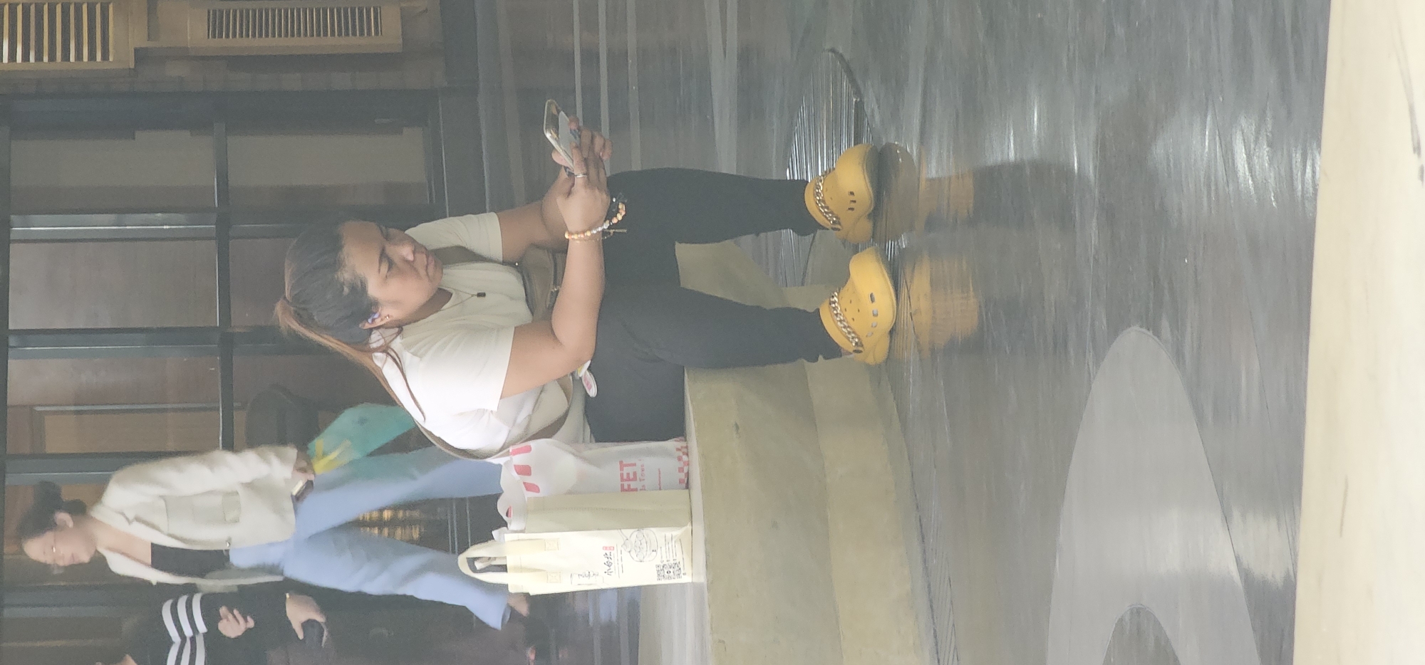 A woman of Asian descent is seated on a concrete ledge, looking down at her mobile phone. She has her hair pulled back in a ponytail and is wearing a white t-shirt, black leggings, and yellow Crocs with a gold chain embellishment. A brown crossbody bag is slung over her shoulder. Next to her on the ledge are two shopping bags, one beige and one white with red lettering. In the background, another woman in a white blazer and light blue pants is walking away. The setting appears to be an indoor or covered area with large windows and reflective flooring. The scene is likely in Makati City, Philippines, given the context provided.