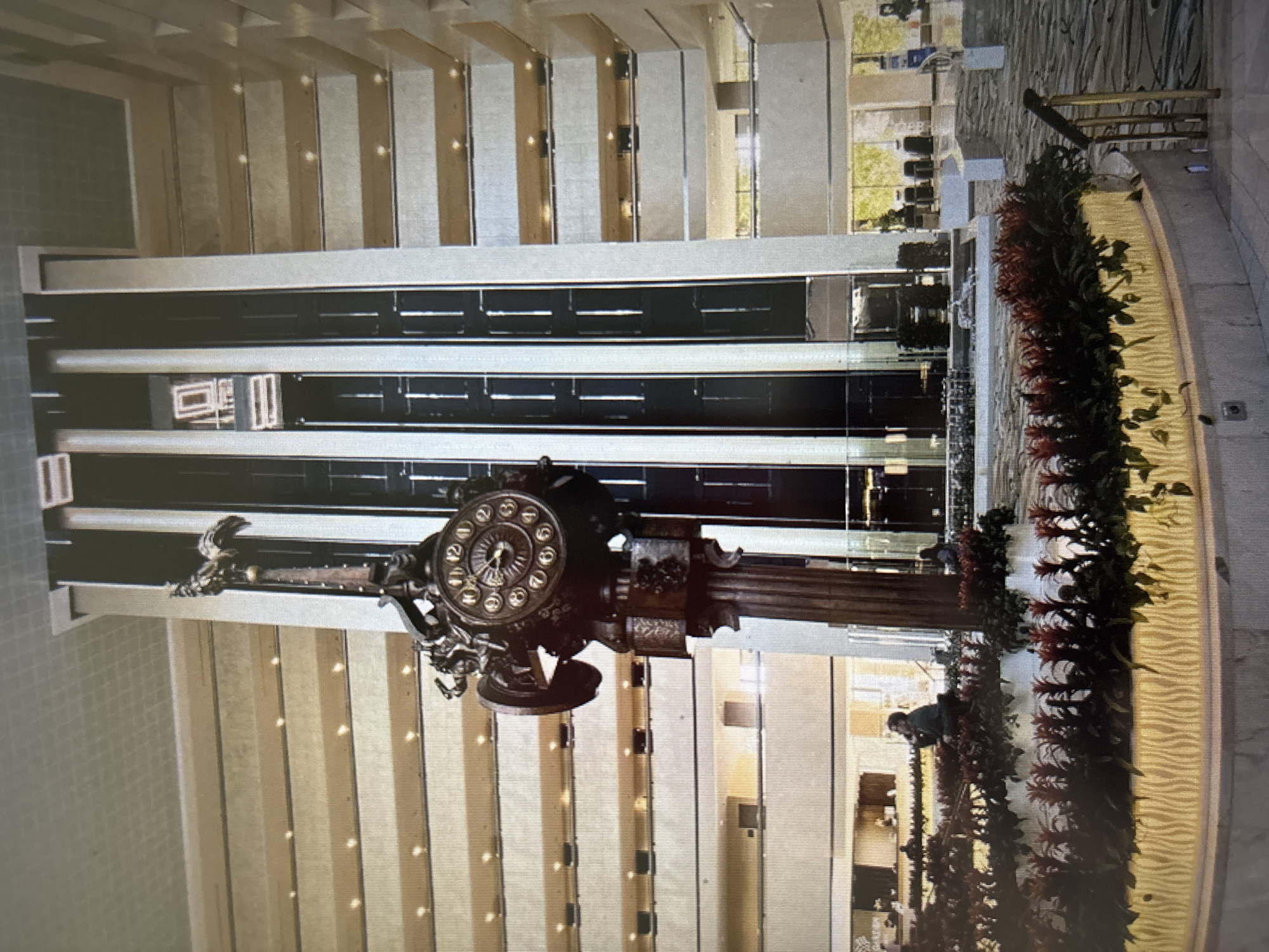 The image shows a multi-story atrium, likely a hotel lobby or convention center, with a large, ornate clock as a central feature. The clock is mounted on a decorative pedestal and appears to be made of dark metal with intricate carvings. The atrium has multiple levels of balconies or walkways with railings, and a modern glass elevator shaft with vertical black panels. In the foreground, there is a circular planter filled with red and green foliage, illuminated from below. Several people are visible in the atrium: one person is sitting on the edge of the planter, looking towards the elevator, while another person is further in the background, near a counter. The overall environment appears to be indoors, with artificial lighting and a sense of spaciousness. The location is not specified.
