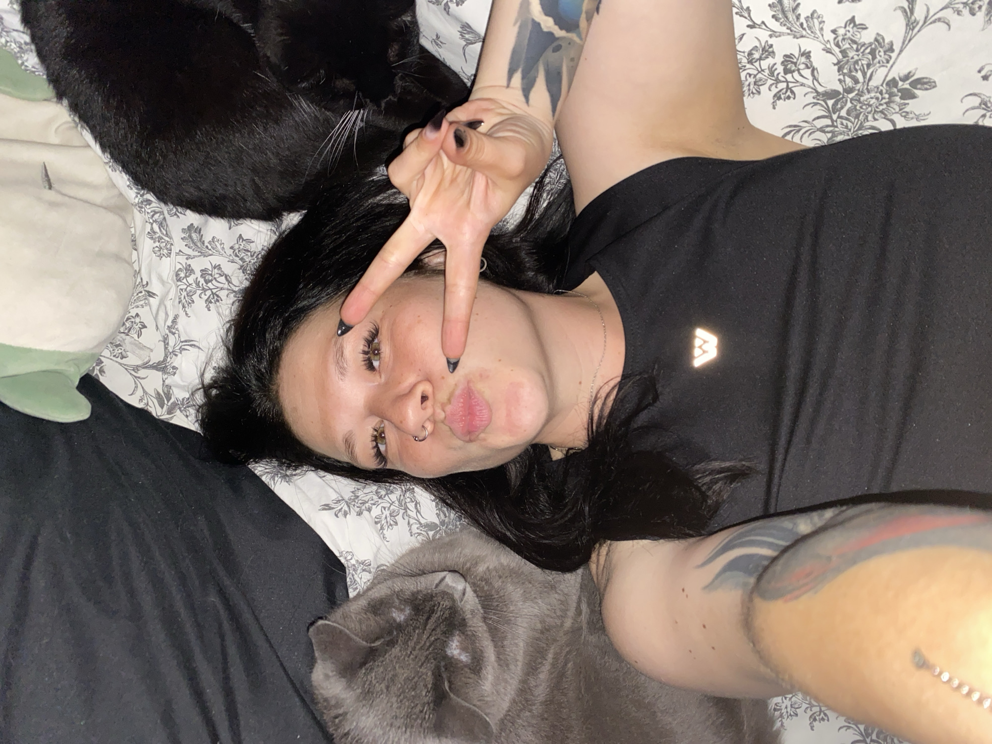A woman with dark hair and multiple tattoos is lying on a bed with two cats. One cat, a grey British Shorthair, is curled up to her left. A black cat is partially visible behind her head and to her right. The woman is wearing a black tank top with a white "W" logo. She is making a peace sign with her right hand and puckering her lips. The bed is covered with a white sheet with a floral pattern. The background is dark, suggesting the photo was taken at night or in low light conditions. The location is not specified in the image.