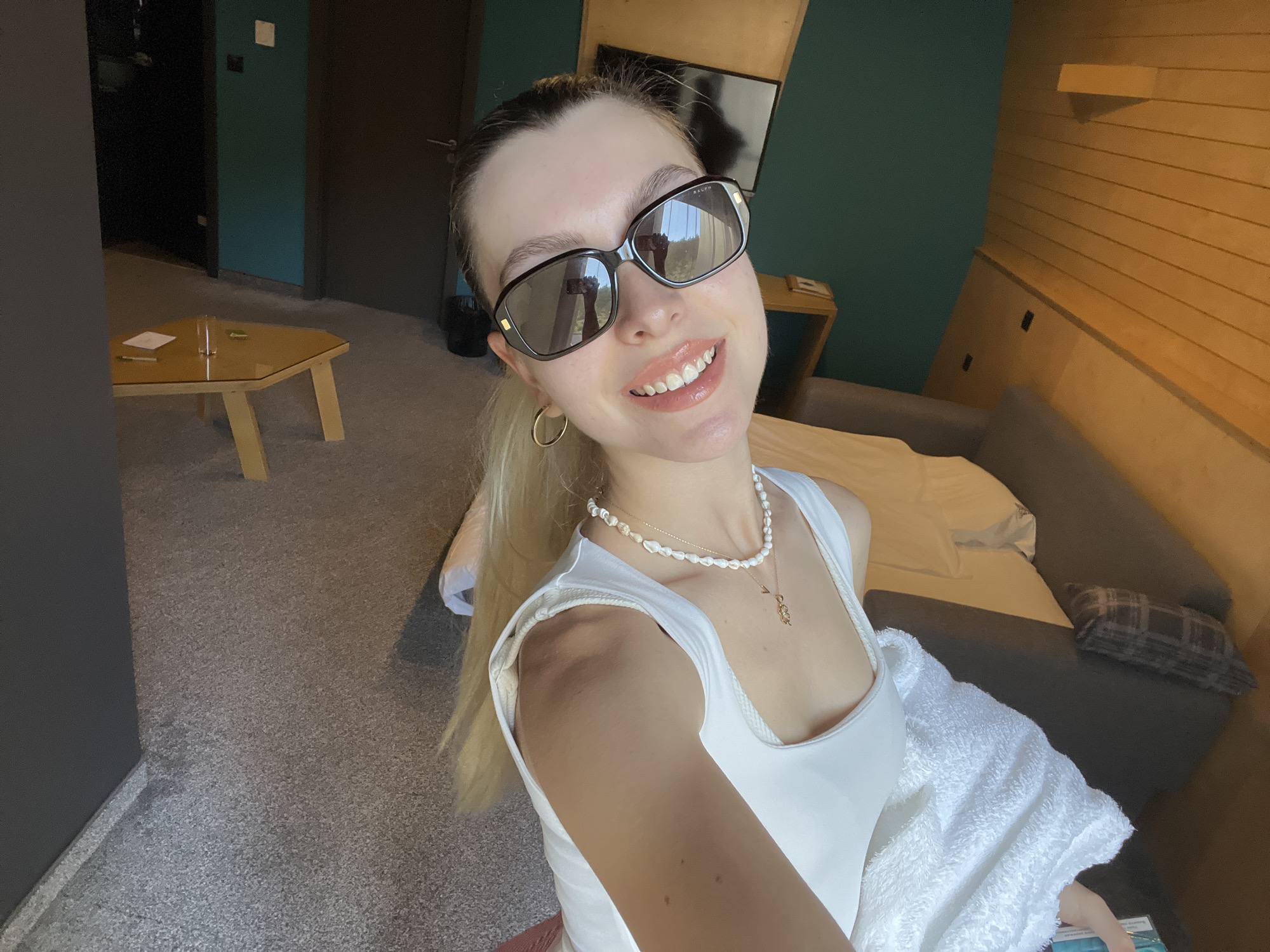 A young woman is taking a selfie in what appears to be a hotel room. She is wearing sunglasses, a white tank top, a pearl necklace, and hoop earrings. She has light blonde hair pulled back in a ponytail. Behind her, there is a bed with gray and white pillows and bedding, a wooden wall, and a side table. In the background, there is a small table with objects on top of it. The walls are painted in teal and gray. The floor is covered in gray carpet. The specific location and city are unknown.