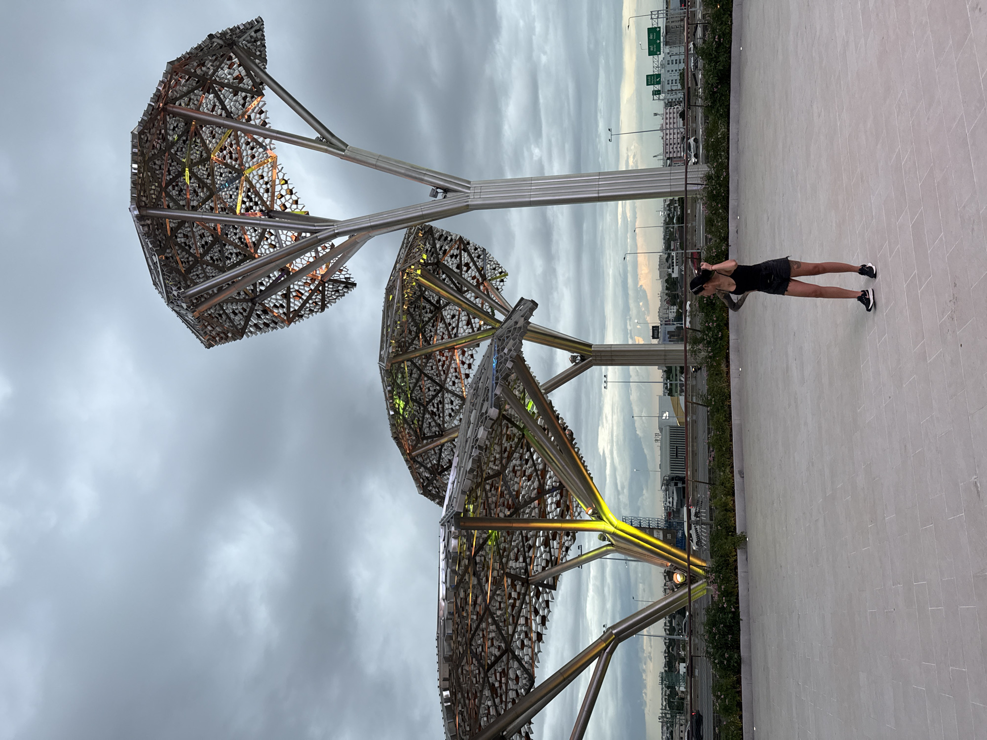 In Bang Kaeo Town Municipality, Thailand, a woman with dark hair and tattoos stands on a light-gray tiled plaza, looking to her right. She is wearing a black tank top and black denim shorts, along with black and white athletic shoes. In the background, three large, stylized metallic trees with hexagonal canopies rise against a cloudy sky. The trees are illuminated with warm yellow and green lights, visible through the intricate metalwork of their canopies. Beyond the trees, a highway with traffic and city buildings can be seen. The overall atmosphere suggests an evening scene in an urban environment with modern architectural installations.