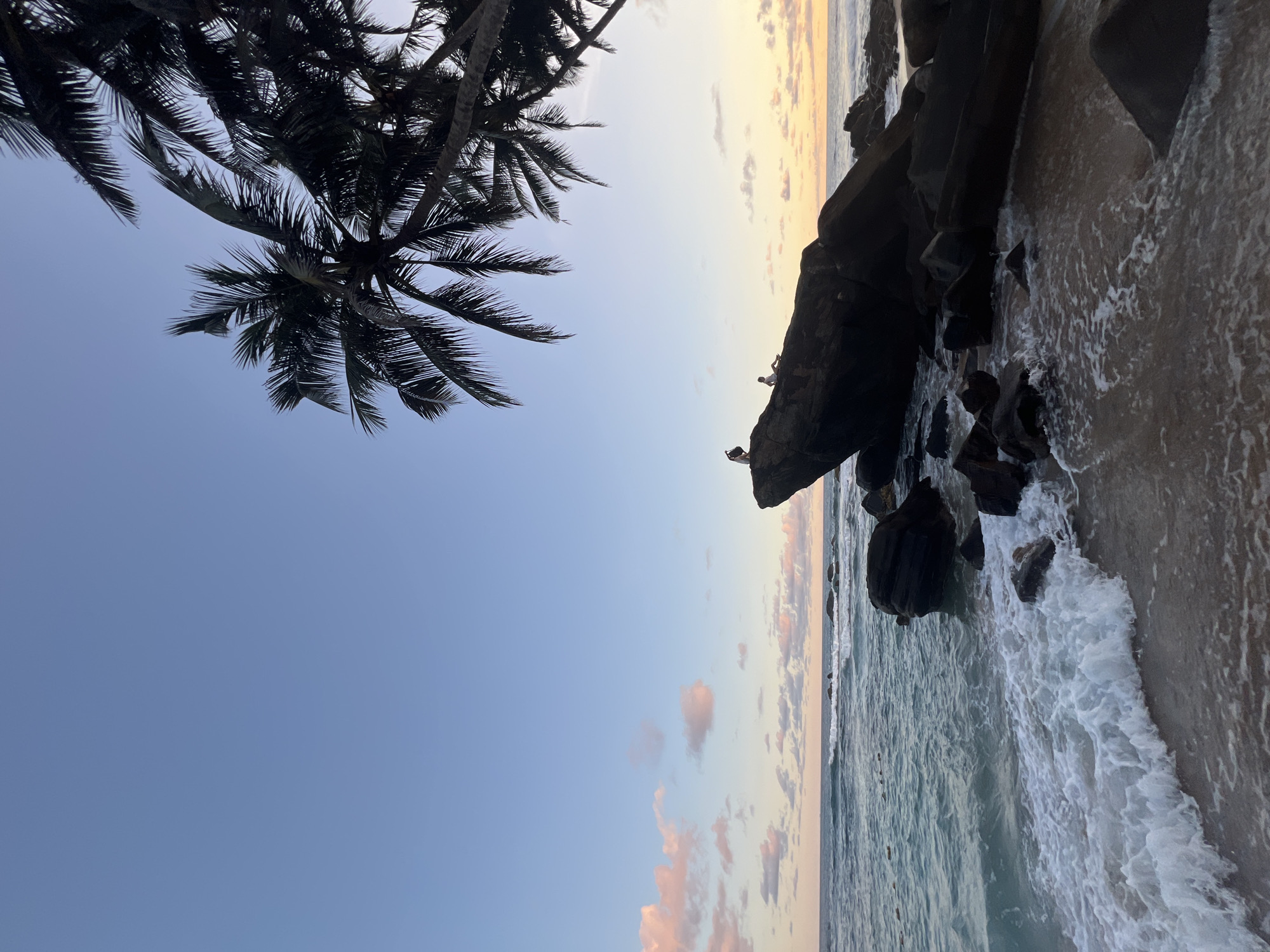 The image captures a beach scene in Talpe, Sri Lanka, during what appears to be sunrise or sunset. The sky transitions from a clear blue to shades of pink and yellow towards the horizon. The ocean is present, with waves gently lapping against the shore. Dark rocks are scattered along the shoreline, against which the waves are crashing. Two small figures, likely people, are positioned on top of the rocks, appearing as dark silhouettes. Silhouetted palm trees are visible at the top left of the frame, adding a tropical feel to the scene.