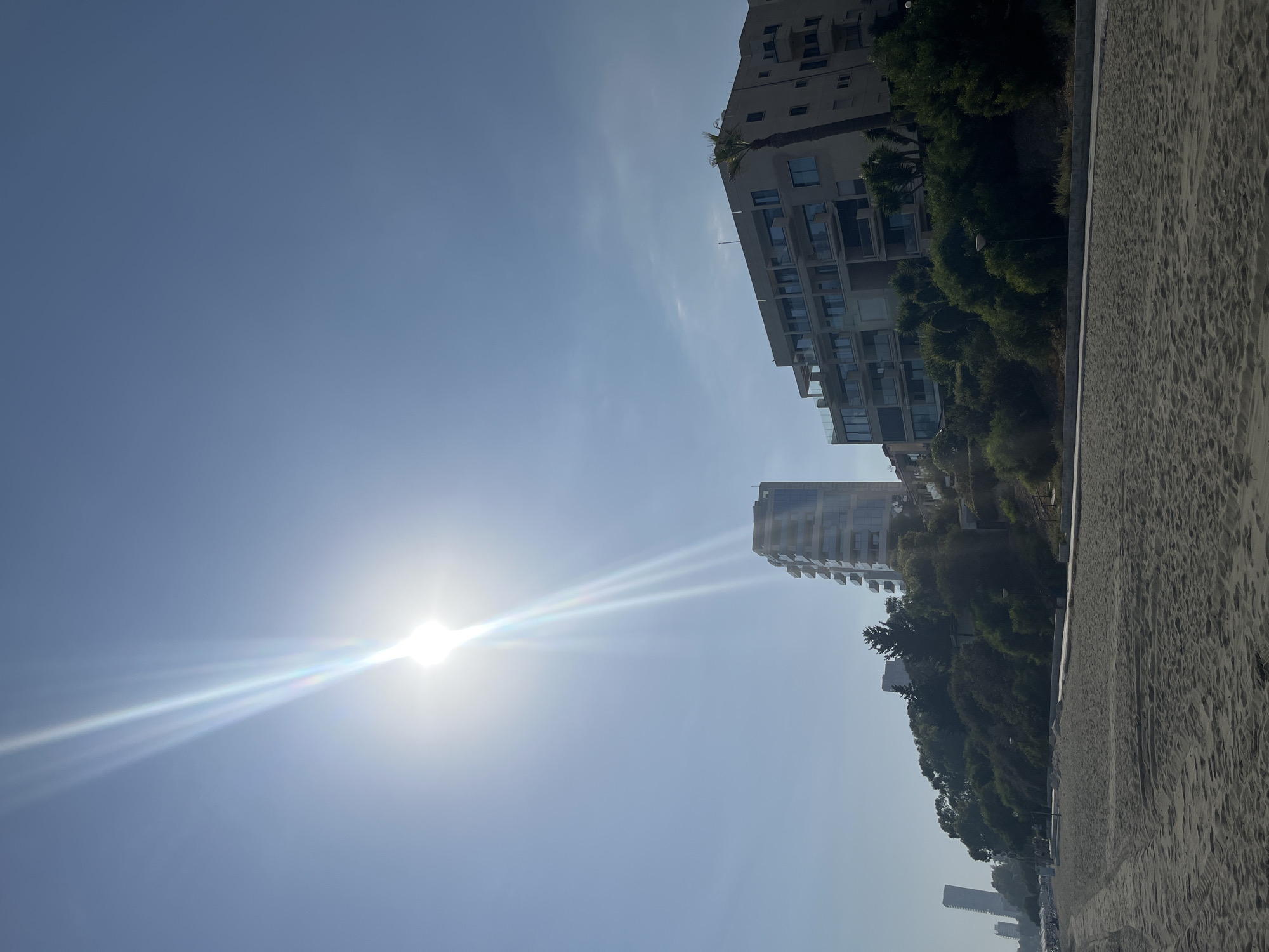 The image captures a bright, sunny day at a sandy beach in Agios Tychona Community, Cyprus. The sun is high in the sky, causing a strong lens flare that radiates downwards. In the background, several modern buildings are visible, some are tall and slender, while others are wider and tiered. These buildings are situated on a vegetated hillside overlooking the beach. The beach itself is covered in light-colored sand, with tire tracks from a vehicle visible in the foreground. A paved pathway runs along the edge of the beach, bordered by lush green bushes and trees. A palm tree is visible next to one of the buildings. The sky is a clear, vibrant blue, with a few faint clouds. No people or specific actions are discernible in the scene.