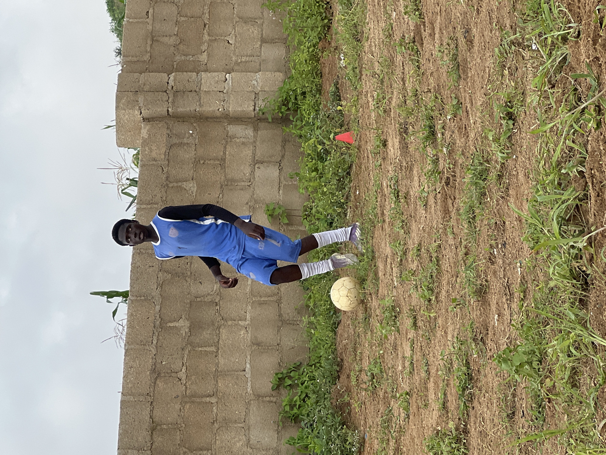 A young Black male is dribbling a soccer ball on a dirt field in Jos, Nigeria. He is wearing a blue soccer jersey with white trim and a blue soccer shorts, a black long-sleeved shirt underneath the jersey, white socks, and athletic shoes. He is looking down at the ball, which is at his feet. In the background, there is a brick wall with green vegetation growing around it. The sky is overcast. There is a small orange traffic cone visible on the right side of the image. The scene appears to be an outdoor soccer practice or casual game.