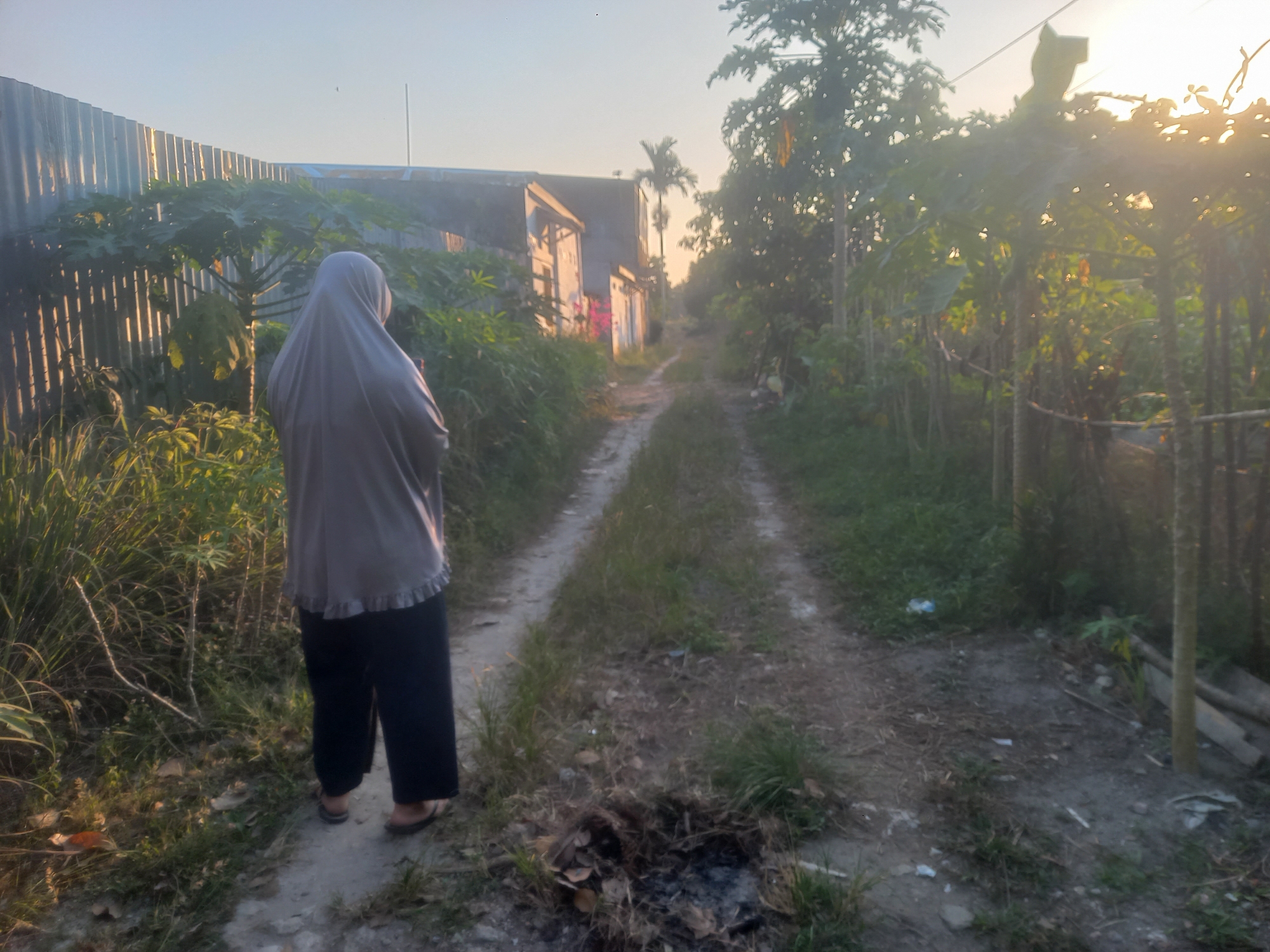 An individual wearing a grey hijab and dark pants stands facing away from the camera on a dirt path. The path leads into the distance between overgrown vegetation and a weathered building on the left. The building appears to be made of corrugated metal and wood, with some graffiti visible on its side. The sun is setting or rising, casting a warm glow over the scene and creating lens flare in the upper right corner. The environment appears rural, consistent with a location in Kasang Pudak, Indonesia.