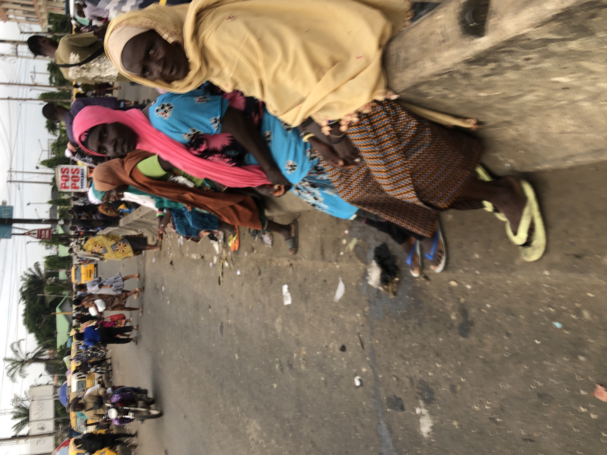 The image depicts a street scene in Ikeja, Nigeria, with a focus on several women sitting on a concrete barrier along the roadside. The women are dressed in brightly colored clothing and headscarves, suggesting a cultural context. In the background, a busy street with traffic, including motorcycles and cars, is visible, indicating an urban environment. There are signs and buildings in the distance, further confirming the city setting. The ground is littered with debris, suggesting a lack of cleanliness in the area. The overall impression is one of everyday life in a bustling Nigerian city.