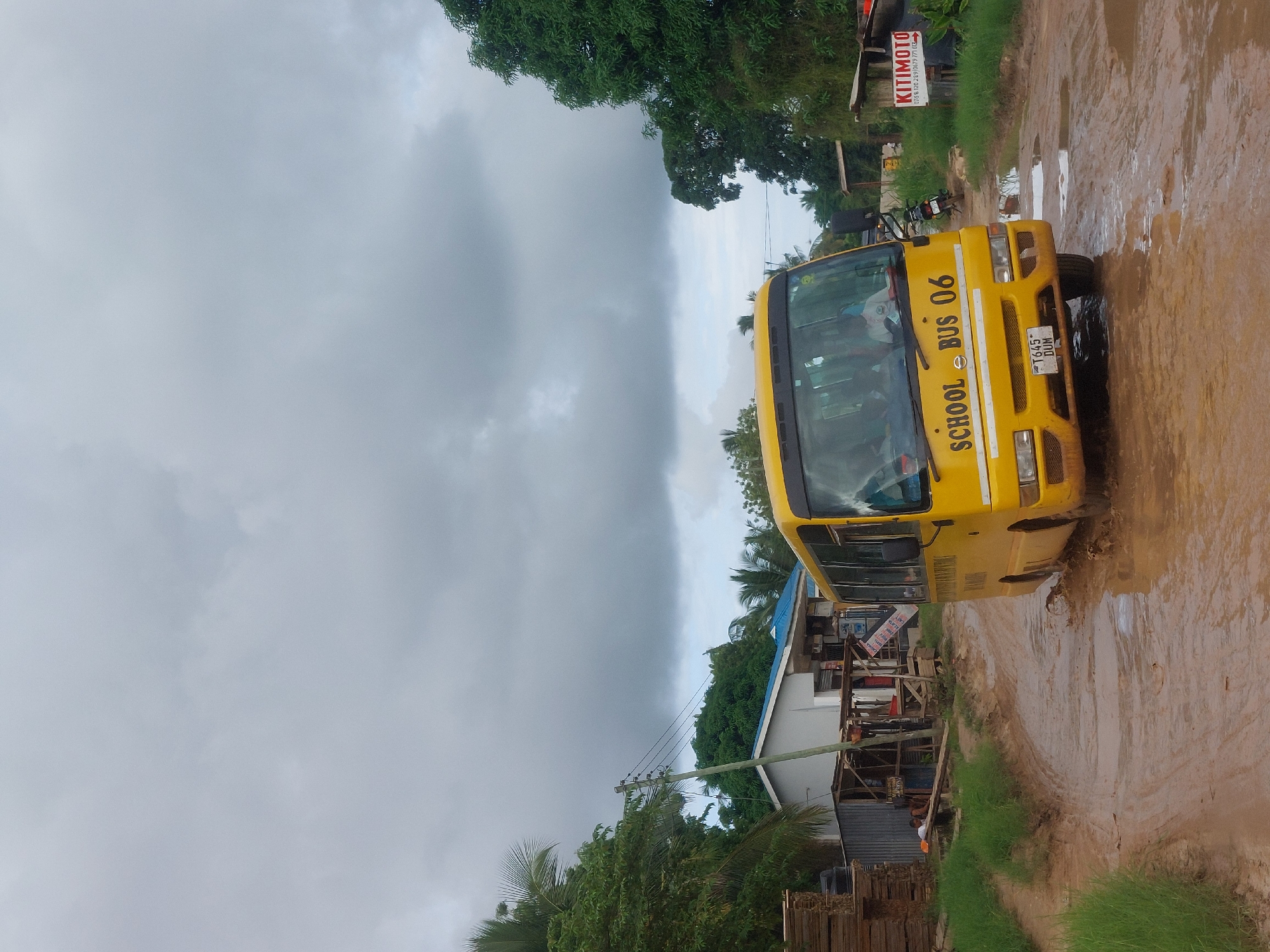 A yellow "SCHOOL BUS 06" is driving on a muddy road in what appears to be a residential area. The license plate reads "T 645 DOM." There are buildings and vegetation on either side of the road. A sign with "KITIMOTO" is visible. The sky is overcast. Based on the sign and the license plate style, the location is likely in or near Dar es-Salaam, Tanzania.