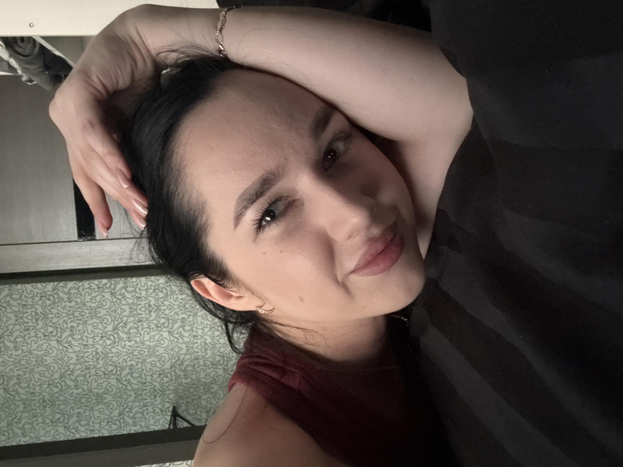 A young woman with dark hair lies on a bed in Novosibirsk, Russia. She is wearing a burgundy tank top and a delicate bracelet on her right wrist. Her left hand is resting on her head, and her head is tilted to the side, supporting her head on her arm. The background shows a patterned wallpaper and part of a closet. The lighting is soft, creating a relaxed and intimate atmosphere.
