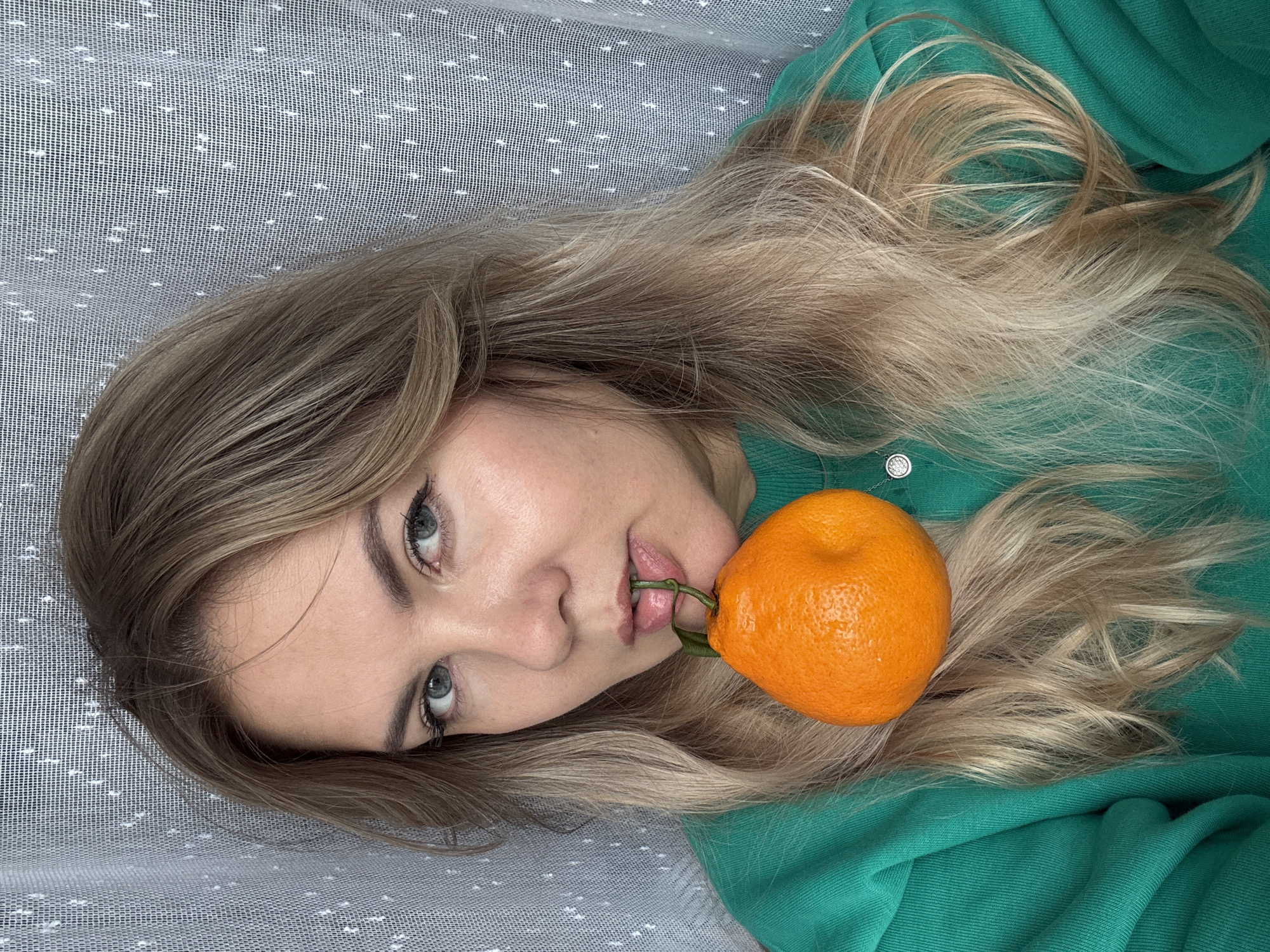 A woman with blonde hair, wearing a green sweatshirt, holds an orange in her mouth. The background consists of a white, translucent curtain with a dotted pattern. The image appears to be taken indoors, possibly in an apartment in Izhevsk, Russia. The woman's face is partially visible, and she is looking directly at the camera. The focus is on her face and the orange.