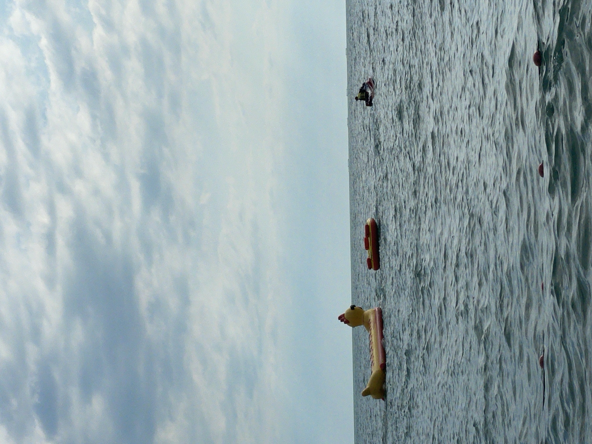The image shows a body of water with a cloudy sky above. On the water, there are several objects, including inflatable toys and a person on a board. In the foreground, a yellow inflatable toy resembling a giraffe is partially submerged. Further in the distance, a red and yellow inflatable toy shaped like a hot dog floats on the water. There is also a person on a small board, appearing to be wakeboarding or water skiing, with an American flag attached to their equipment. The water is choppy, indicating it may be windy or there are waves. The overall scene suggests an outdoor recreational activity taking place on a lake or the sea. The weather appears overcast, with a light blue sky filled with soft, wispy clouds. The location is identified as Sochi, Russia. The time of day appears to be daytime, given the lighting conditions. There is no visible text in the image.