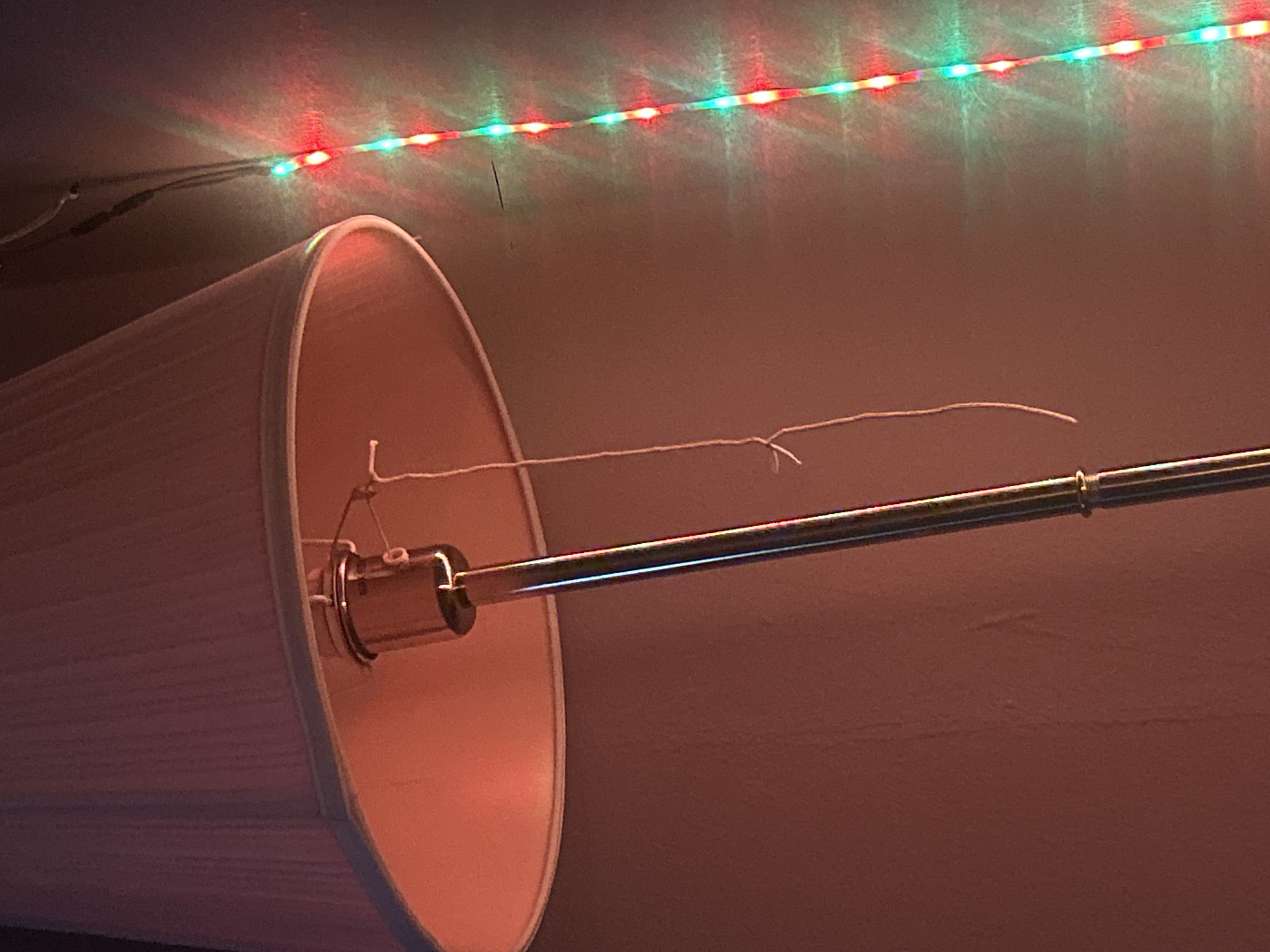 The image shows a lamp with a white shade, suspended by a metal rod, set against a plain wall. A string or cord is attached to the lampshade, extending out towards the right. Above the lamp, a strand of red and green LED lights is affixed to the ceiling. The environment appears to be indoors, possibly a room with warm-toned lighting. There is no information in the image that indicates the location is Kalitinovo, Bulgaria.