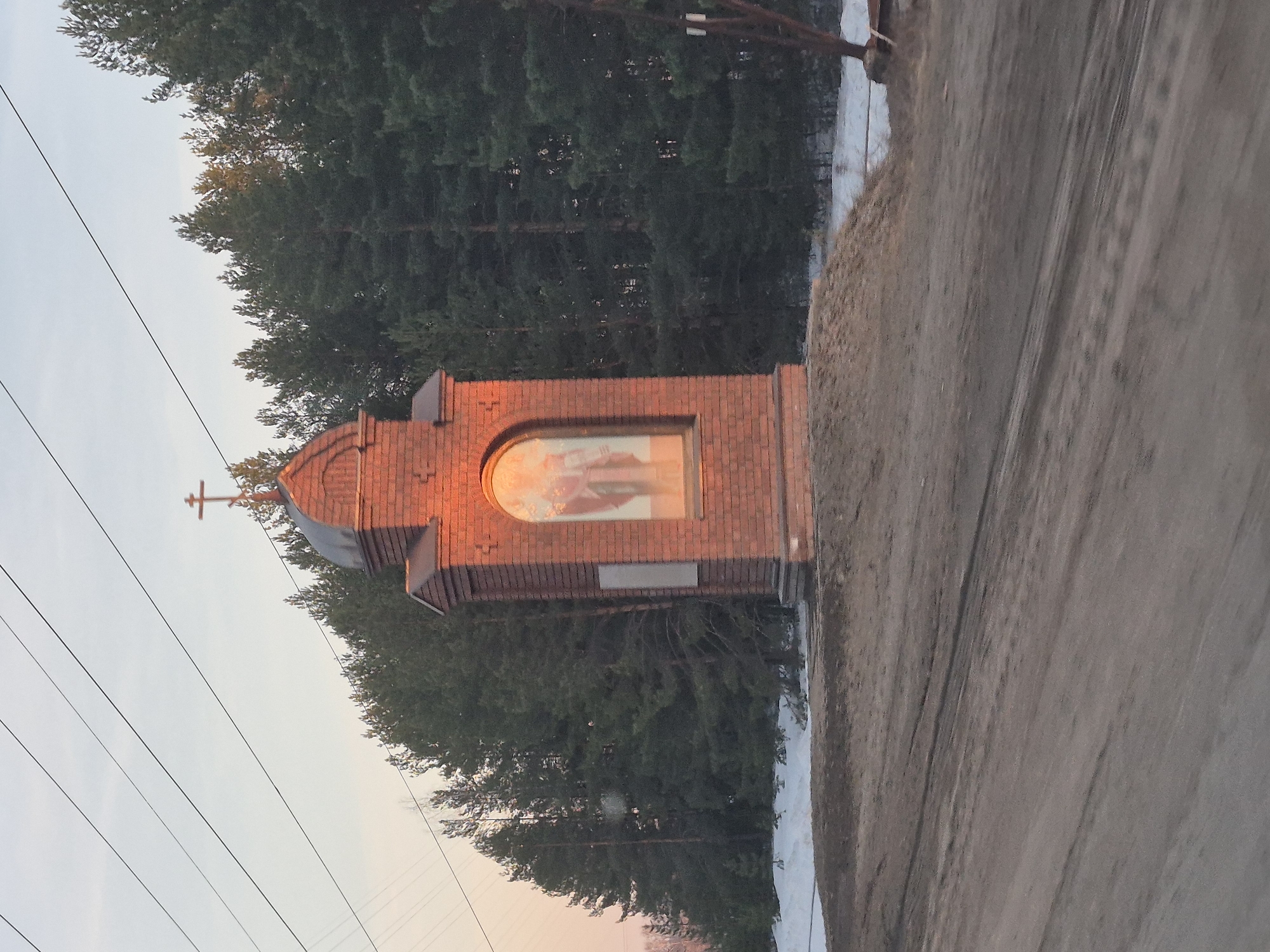 The image shows a brick wayside shrine in Noviy, Russia. The shrine has a cross on top and a painting inside an arched window. It stands beside a dirt road with some snow on the verge. A dense forest is behind the shrine, and power lines are visible in the sky.