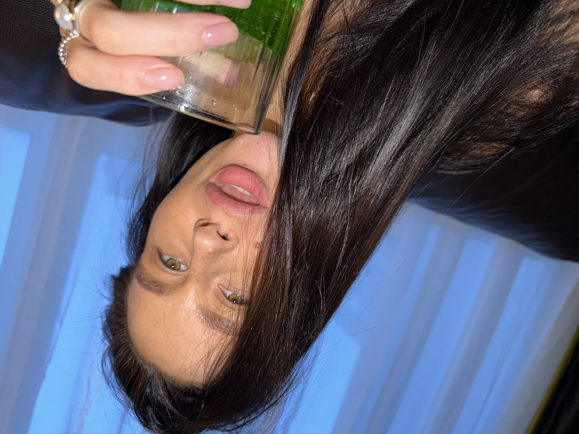 The image is a close-up, rotated 90 degrees counter-clockwise, featuring a person indoors. The central subject is a person, likely female, with fair skin, dark long hair, and prominent green eyes. Her lips are adorned with pink lipstick and are slightly parted, revealing her teeth. Her dark, straight hair cascades around her face and fills a significant portion of the frame.
In the upper right section of the image, a hand is visible, holding a clear glass filled with a bright green liquid. The hand has neatly manicured, oval-shaped nails with a light pink polish. Two rings are worn on the fingers: one silver-toned ring with a large, light-colored gemstone (possibly a pearl or moonstone) surrounded by smaller stones, and another silver-toned ring with a textured or braided design. The hand and part of a dark-colored surface, possibly clothing or furniture, are visible.
The background primarily consists of a light blue area with distinct vertical lines, strongly suggesting window blinds or curtains. This indicates an indoor setting, possibly near a window, in Khimki, Russia. The lighting appears soft and even, highlighting the subject's features. The person's expression is neutral, with her mouth slightly open, appearing to be in the process of holding or drinking from the glass. There is no visible text in the image.