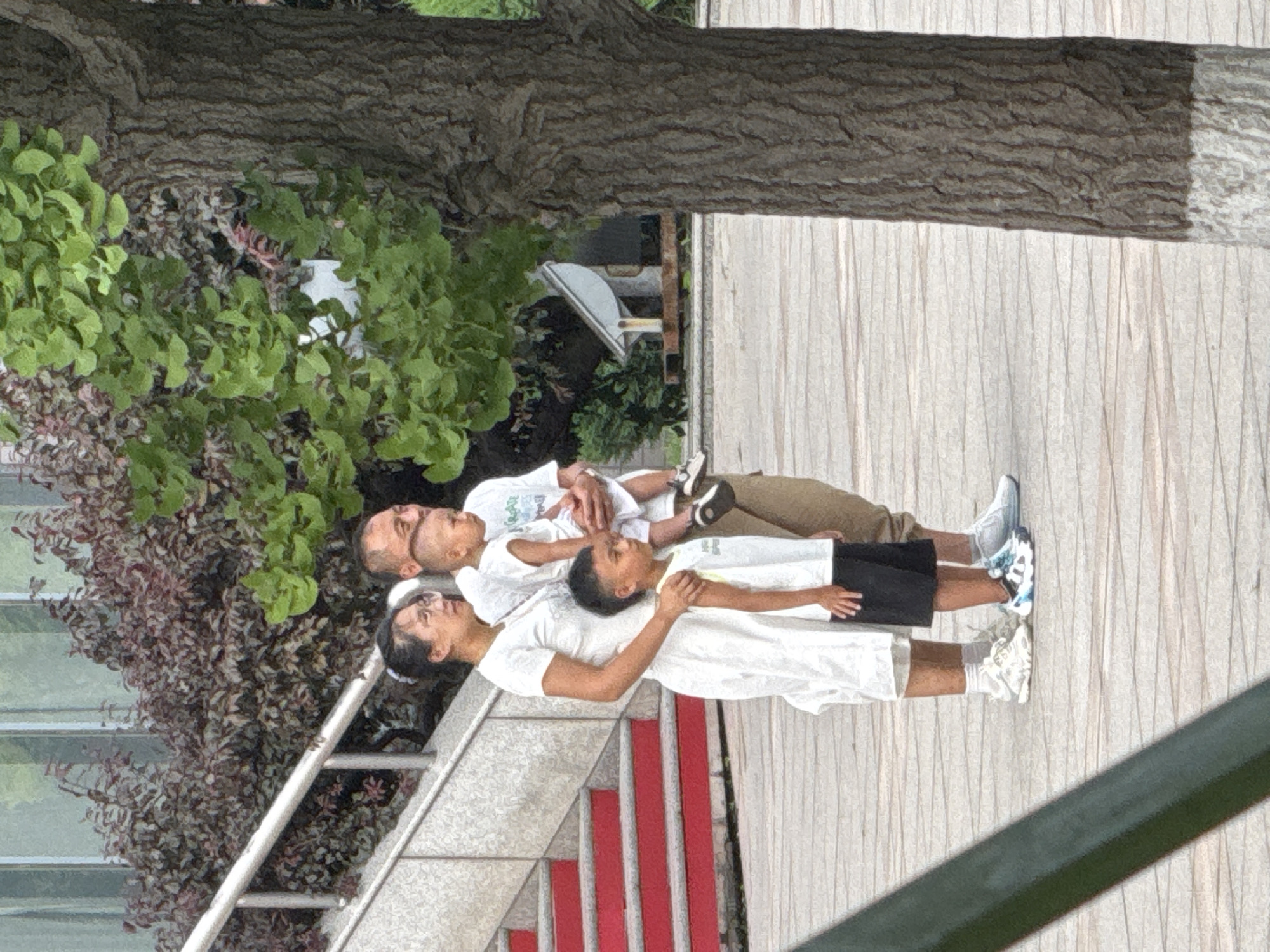 A family of four, consisting of two adults and two young boys, stands on a paved area near a staircase with red steps and metal railings. The family is positioned in front of a large tree and a cluster of trees with red and green foliage. The father, in the middle, holds a baby in his arms, while the mother stands to his left, looking forward. The older son, wearing a white tank top and black shorts, stands to the left of the father, facing to the right. The family is dressed casually in white tops and light-colored pants or shorts. The background shows a building with large windows and some distant structures. The lighting suggests it is daytime.
