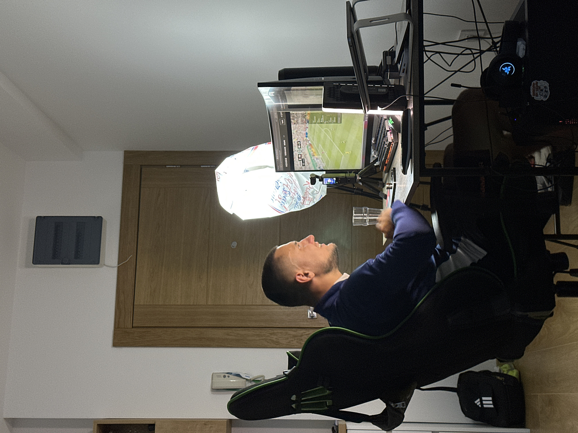 A medium shot captures a man sitting in a gaming chair, facing a computer setup in what appears to be a residential room in Donji Pobori, Montenegro. The man, with a buzz cut and wearing a dark blue jacket over a white shirt, is looking to his right, with his mouth slightly open as if speaking or in mid-action. His hands are resting on the desk.
The desk is cluttered with electronic devices. Two computer monitors are prominent; the left one displays a soccer video game, while the right one is mostly black. A keyboard, mouse, and a small camera on a tripod are also on the desk. A tall, diffused lighting fixture, shaped like a sphere and covered in white fabric, is positioned behind the left monitor, suggesting a streaming or recording setup. A glass of water sits on the desk to the man's left.
To the left of the man, the back and armrest of a black and green gaming chair are visible. A black Adidas backpack with white stripes is hung on the back of the chair. In the background, a light brown wooden door stands closed. Above the door and slightly to the left, a dark gray electrical panel is mounted on a white wall. The room's flooring is a light wood laminate. The overall lighting is dim, with the computer screens and the lighting fixture being the primary light sources.