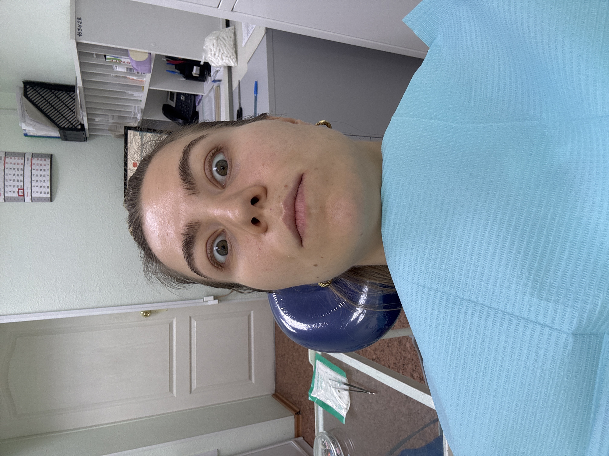 A medium shot depicts a person lying back in a dental chair, covered with a light blue dental bib. The person has fair skin, brown hair, and brown eyes, looking directly at the camera with a slightly apprehensive expression. They are wearing small gold hoop earrings. The headrest of the dental chair is visible in dark blue behind the person's head.
In the background, a white door is visible on the left, with a gold-colored door handle. To the left of the person's head are a calendar and a filing system, as well as a desk.