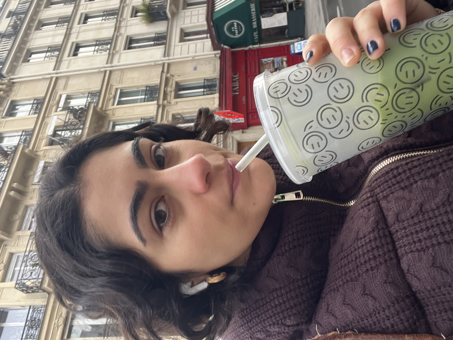 A person with dark, wavy hair, wearing a dark plum-colored textured puffer jacket, is depicted taking a selfie while drinking from a clear plastic cup with a straw. The individual's left ear features a white wireless earbud and a gold-colored hoop earring. The clear cup is decorated with numerous black outline smiley faces and contains a light green liquid. The person's left hand, with dark nail polish, holds the cup.
The background presents a typical Parisian street scene. Dominant are Haussmannian-style buildings, characterized by their light-colored stone facades, multiple stories, ornate black iron balconies, and tall windows. At street level, a red storefront with "TABAC" in white capital letters is visible. Adjacent to it is a establishment with a dark green awning bearing the text "LA PARISIENNE CAFE - BRASSERIE". The scene is consistent with an urban environment in Paris, France, likely on an overcast day.