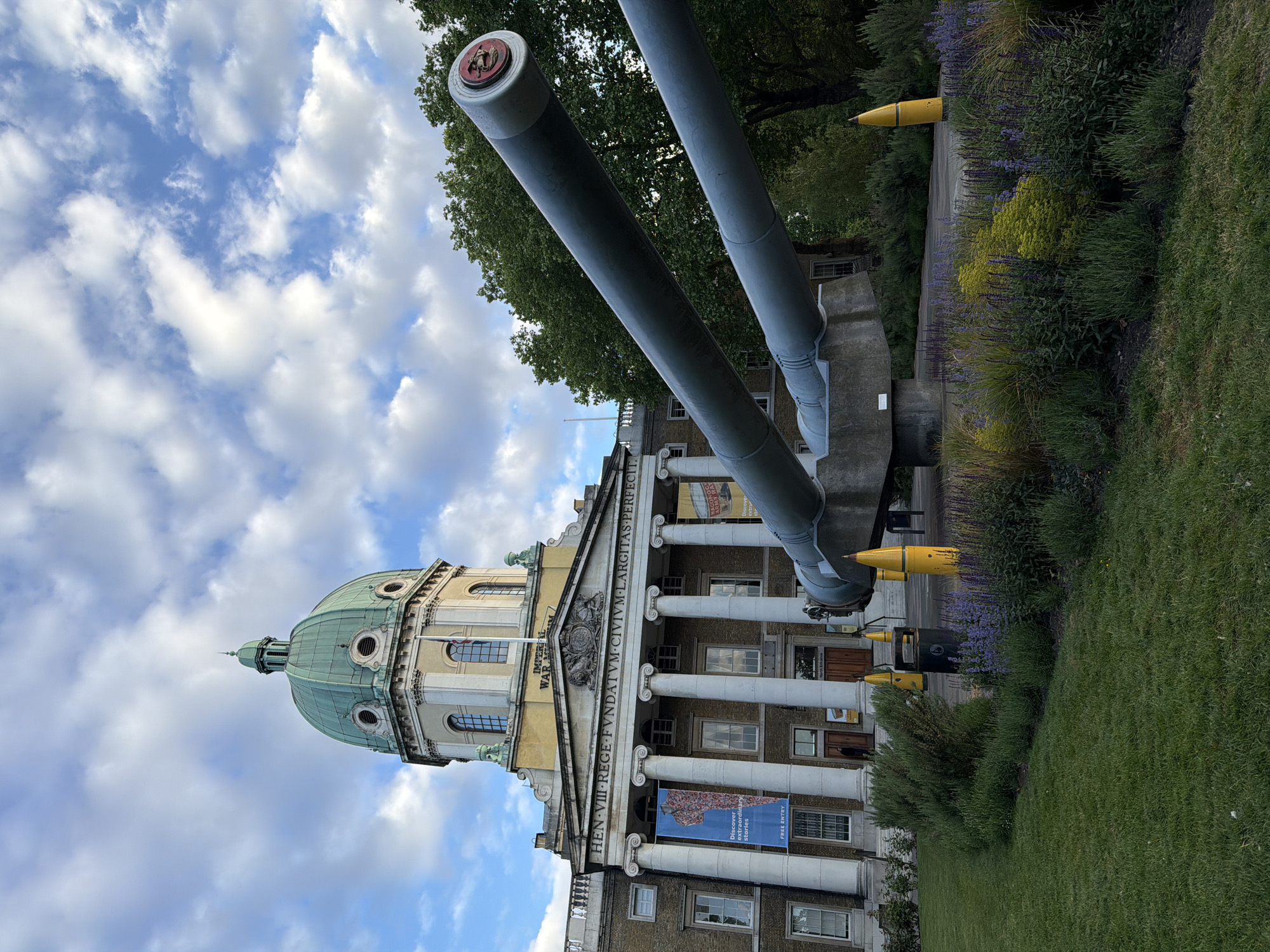 The image depicts the Imperial War Museum in London, United Kingdom. The building is a classical-style structure with a large dome on top and a row of columns at the entrance. Two large naval guns are mounted outside the museum, pointing towards the sky. Several yellow munitions are placed near the guns, and vegetation is on the right-hand side. The sky is partially cloudy.