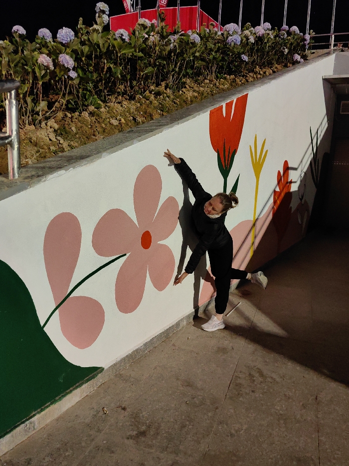 The image captures a nighttime scene in Da Lat, Vietnam, depicting a single individual posing against a white concrete wall adorned with a colorful floral mural. The wall is illuminated by an artificial light source from the left, casting distinct, elongated shadows of the person and elements onto its surface.
Above the wall, a raised garden bed features numerous light purple hydrangea plants and green foliage. A silver metal railing is partially visible on the far left, bordering this elevated section. In the dark background, beyond the garden bed, several red rectangular structures or banners are discernible against the night sky. The ground in the foreground is a dark, textured concrete surface, showing minor debris, including what appears to be cigarette butts, near the individual's feet.
The mural on the white wall consists of stylized floral designs: a large pink flower with a red center and green stem on the left, a red tulip-like flower with a green stem, and yellow abstract shapes further to the right.
The individual is an adult female with light skin and hair pulled back. She wears a black long-sleeved jacket, black pants, and white athletic shoes. A white patterned face covering is visible around her chin/neck. She is striking a pose, leaning backward against the mural wall with both arms extended horizontally outwards, palms open. Her left leg is relatively straight, while her right leg is bent at the knee, with the foot raised and pointing left.
