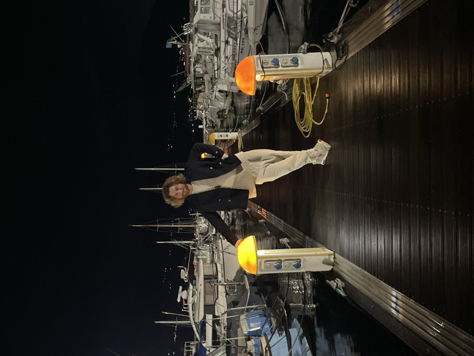 A person in a dark jacket and light pants stands on a dark, wooden boardwalk next to a marina at night. Several boats are docked beside the boardwalk. Yellow lights in white housings are fixed to the boardwalk's edge, providing illumination. The scene has a dark background, highlighting the person and the docked boats.