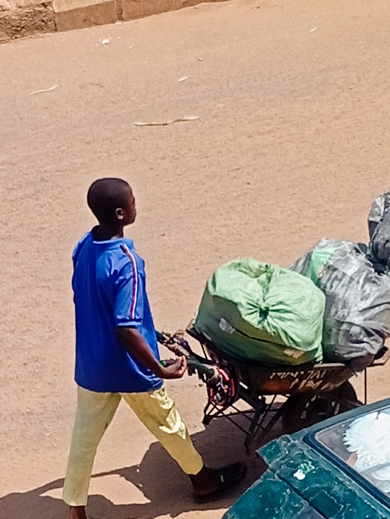 A young Black boy, wearing a blue shirt with red and white stripes on the sleeves and light yellow pants, pushes a wheelbarrow loaded with two large trash bags. One bag is bright green, and the other is gray and appears to be a plastic bag with writing on it. The wheelbarrow has black wheels and a metal frame. The boy is facing to the right of the frame and appears to be in motion. In the foreground, a portion of a dark green vehicle is visible, with its windshield reflecting the sky. The ground is a dirt or gravel surface, common in the city of Bukuru, Nigeria. The scene is set outdoors, under daylight conditions.