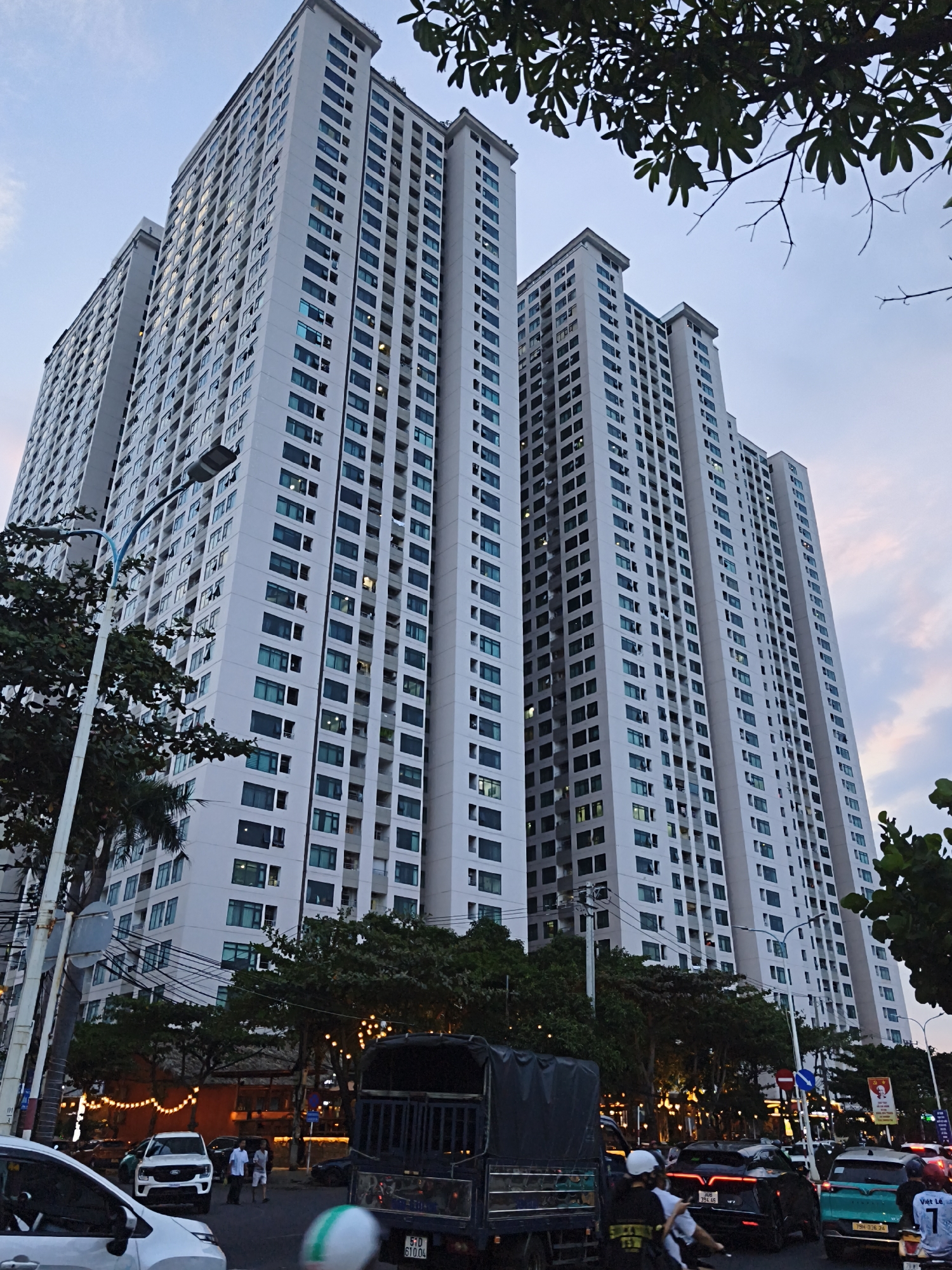 This is a low-angle, medium shot of a cluster of tall, modern apartment buildings in Bac Nha Trang Ward, Vietnam. The buildings are white with many windows, some of which are illuminated. The sky above is a dusky blue with hints of pink, suggesting it's either sunrise or sunset. In the foreground, there's a street with traffic. A white SUV, a black truck, and several motorcycles are visible. There are also a few people on the street, some appearing to be pedestrians and others on motorcycles. Trees line the street in front of the buildings, and some of them are lit with decorative string lights. The overall impression is of an urban environment at dusk.