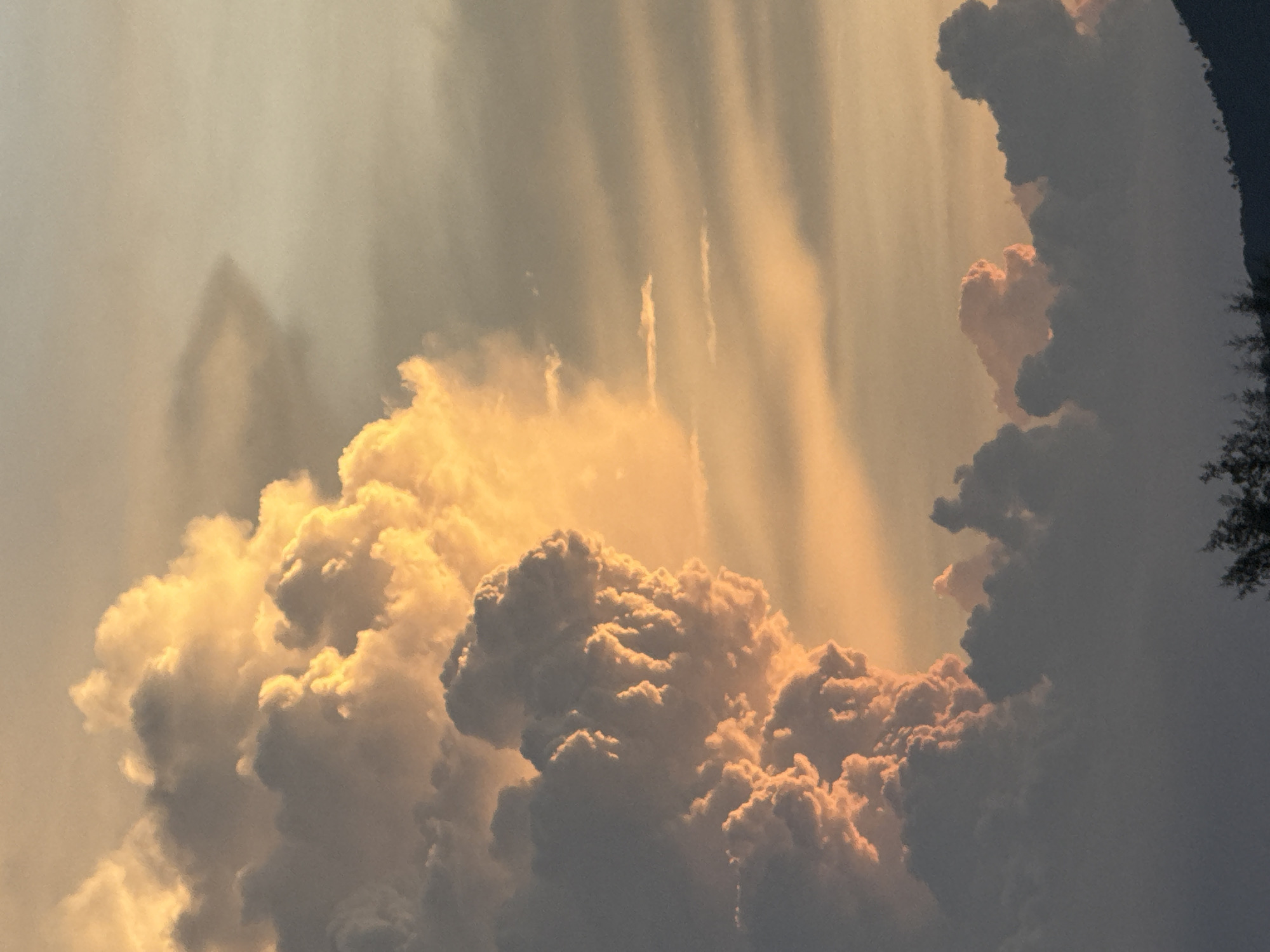 This is a photograph of a dramatic cloud formation at sunset, observed from Baan Hua Thanon, Thailand. The clouds are illuminated from behind by the setting sun, casting a warm golden-orange glow on their upper surfaces, while their undersides are in shadow, appearing in shades of grey and dark blue. The clouds are voluminous and textured, with some resembling towering columns and others forming a more diffused, layered mass. In the foreground at the very bottom of the image, a dark silhouette of trees and a hillside is visible against the brighter sky. The sky itself transitions from a pale yellow near the horizon to a soft greyish-orange overhead, with subtle horizontal cloud streaks visible in the upper right quadrant. There are no people or specific man-made structures clearly discernible in the frame, other than the hint of landmass and vegetation at the bottom.