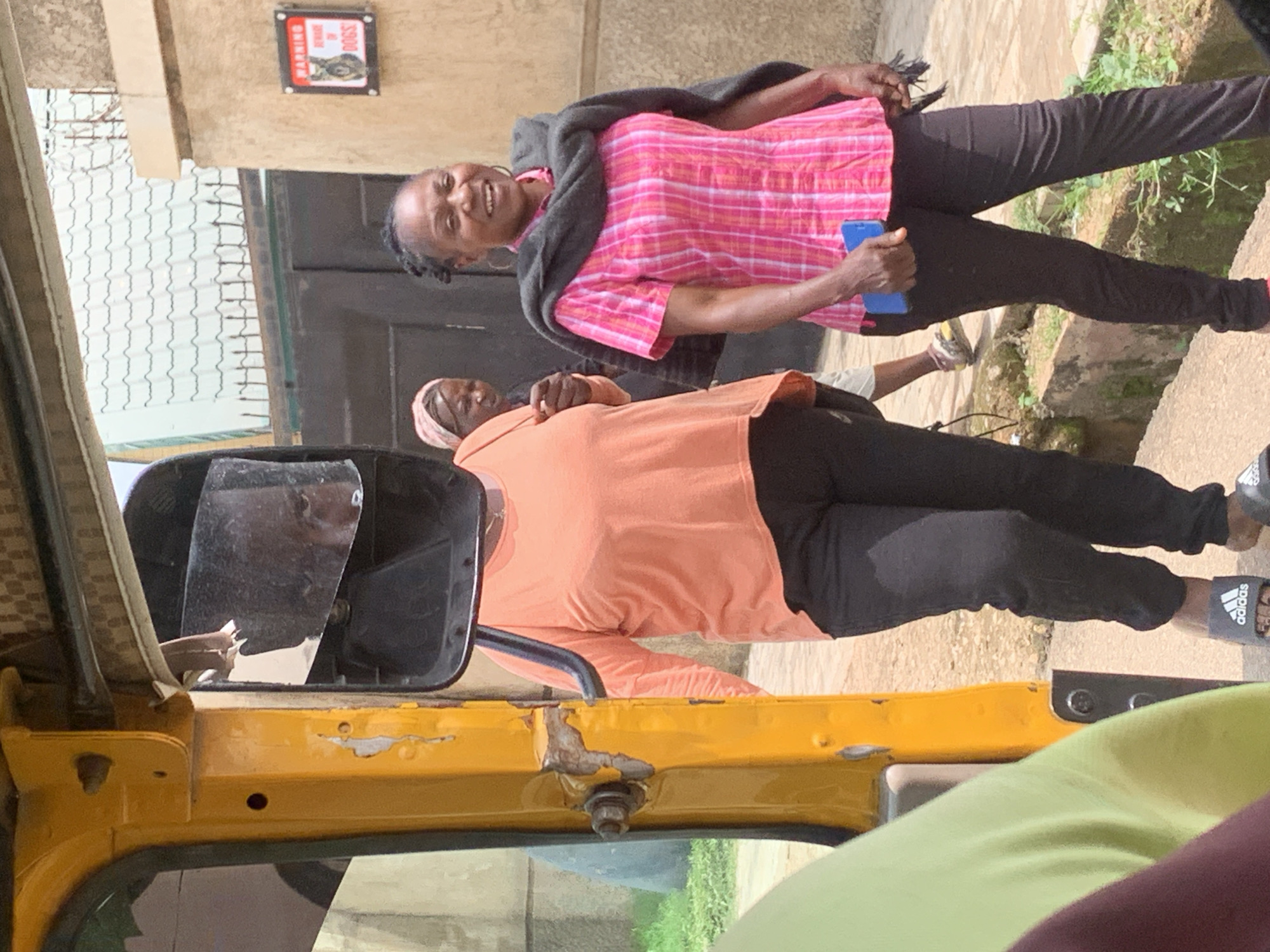 In Jos, Nigeria, two women are seen standing near a yellow vehicle, possibly a tuk-tuk, with one woman in the foreground wearing an orange long-sleeved shirt and black pants. She is looking down and her face is partially obscured. Behind her, another woman is wearing a pink and white checkered shirt and black pants, with a dark gray shawl draped over her shoulders. She is smiling and holding a blue object, possibly a phone. A third person, likely a child, is partially visible behind the woman in the orange shirt, wearing a pink head covering. In the background, there is a wall with a sign that reads "WARNING BEWARE OF DOGS" with a picture of a dog. The scene appears to be outdoors, with hints of greenery and a paved area. The lighting suggests it is daytime.