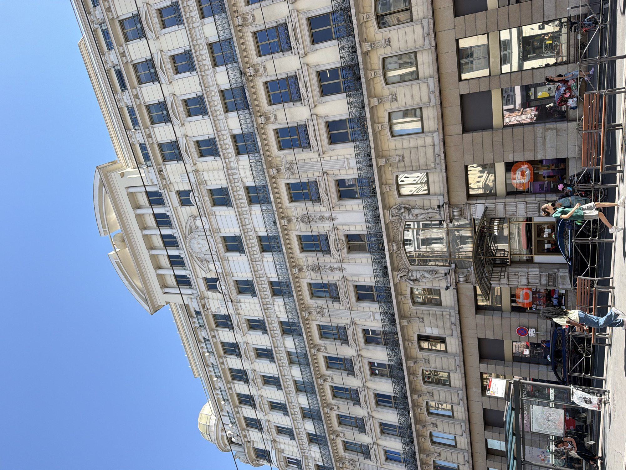 This is a slightly tilted, eye-level shot of a building facade in Lyon, France, on a sunny day. The architecture is detailed, featuring a cream-colored building with multiple rows of windows, each framed by dark metalwork. The building on the right has a more modern, stone-clad facade with large glass windows that reflect the sky and surrounding buildings.
Several people are visible. On the sidewalk, a person wearing a green shirt and shorts stands near a bus stop. Another person, wearing a light-colored shirt and jeans, is leaning against a bench near the bus stop, looking away from the camera. Further down the street, two individuals are seated on a bench in front of the modern building, one wearing a black and white patterned dress and the other a black top. Two bicycles are parked near the building on the right.
The scene appears to be a public area, likely a city street, with a bus stop and benches indicating an urban setting. The sunlight suggests it is daytime, possibly mid-morning or afternoon. The clear blue sky further confirms fair weather. Reflections in the windows of the modern building offer glimpses of other structures and possibly vehicles. There is some visible text on signage, including what appears to be a bus route map inside the bus stop shelter and an orange circular logo on the modern building. The overall mood is casual and ordinary, depicting a typical street scene in a European city.