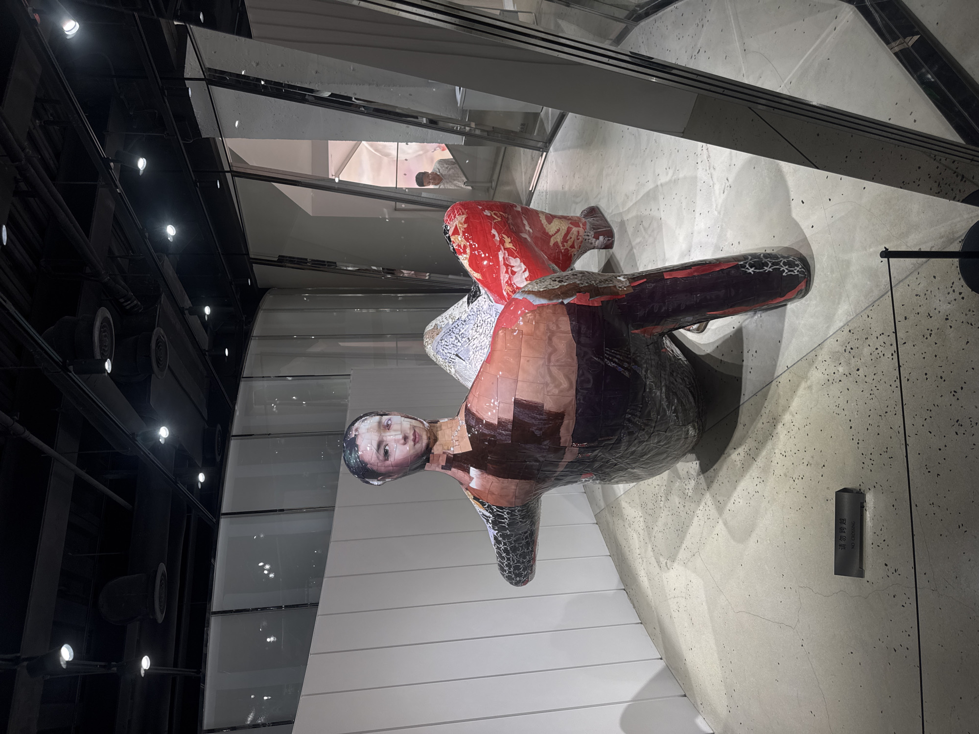 In Shanghai, China, an indoor art exhibition features a large, colorful sculpture resembling a human figure in a seated position. The sculpture is constructed from what appears to be collage elements, with a mosaic-like face of a woman and sections of fabric with intricate patterns, including red with gold dragons and a black and white geometric print. The sculpture is placed on a speckled concrete floor, near a white wall. A small sign on the floor reads "NO CROSSING" in both Chinese and English. In the background, through a glass partition, a man in a light-colored shirt is visible, looking at something within the exhibition space. The environment is brightly lit with track lighting on a dark ceiling grid.
