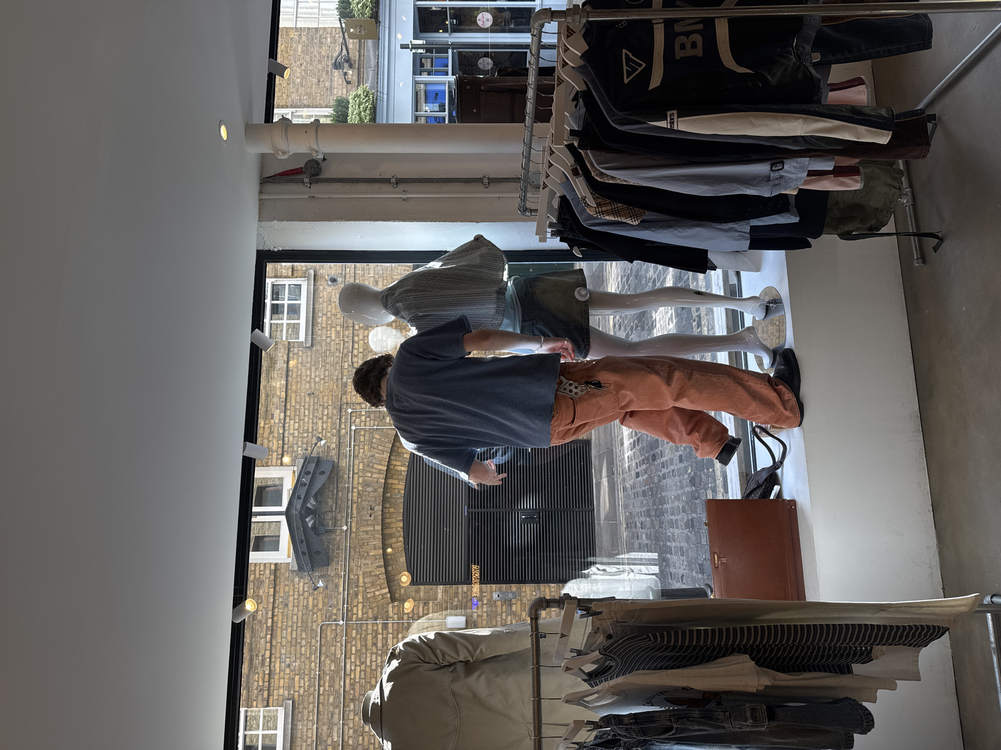 The image depicts the interior of a retail space, possibly a clothing store, in London, United Kingdom. A man wearing a blue shirt and orange trousers stands near a window displaying mannequins. Through the window, the exterior view includes a brick-lined street with an arched entrance marked "ARCHIVE." Various clothing items hang on racks within the store. A brown briefcase is placed on the floor.