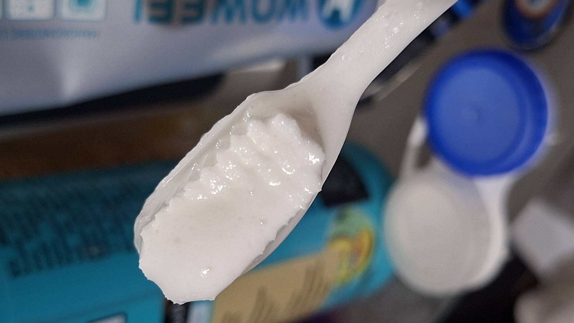 A close-up, shallow depth of field shot focuses on a white, textured paste being held on a white, spoon-like object. The paste appears thick and slightly clumpy, with small glistening beads of moisture visible on its surface. In the blurred background, a bright teal bottle or container with text is partially visible on the left, and a white tube with blue lettering, reading "WOWFEI", is visible on the right. Below and to the right of the paste-laden object, two out-of-focus circular objects are discernible: one is a white lid, and the other is a vibrant blue lid. The overall impression is that of a personal care product, possibly toothpaste or a cosmetic cream, being dispensed or examined in a domestic setting. The image does not contain any elements that allow for specific location identification within Забалоцкі сельскі Савет, Belarus.