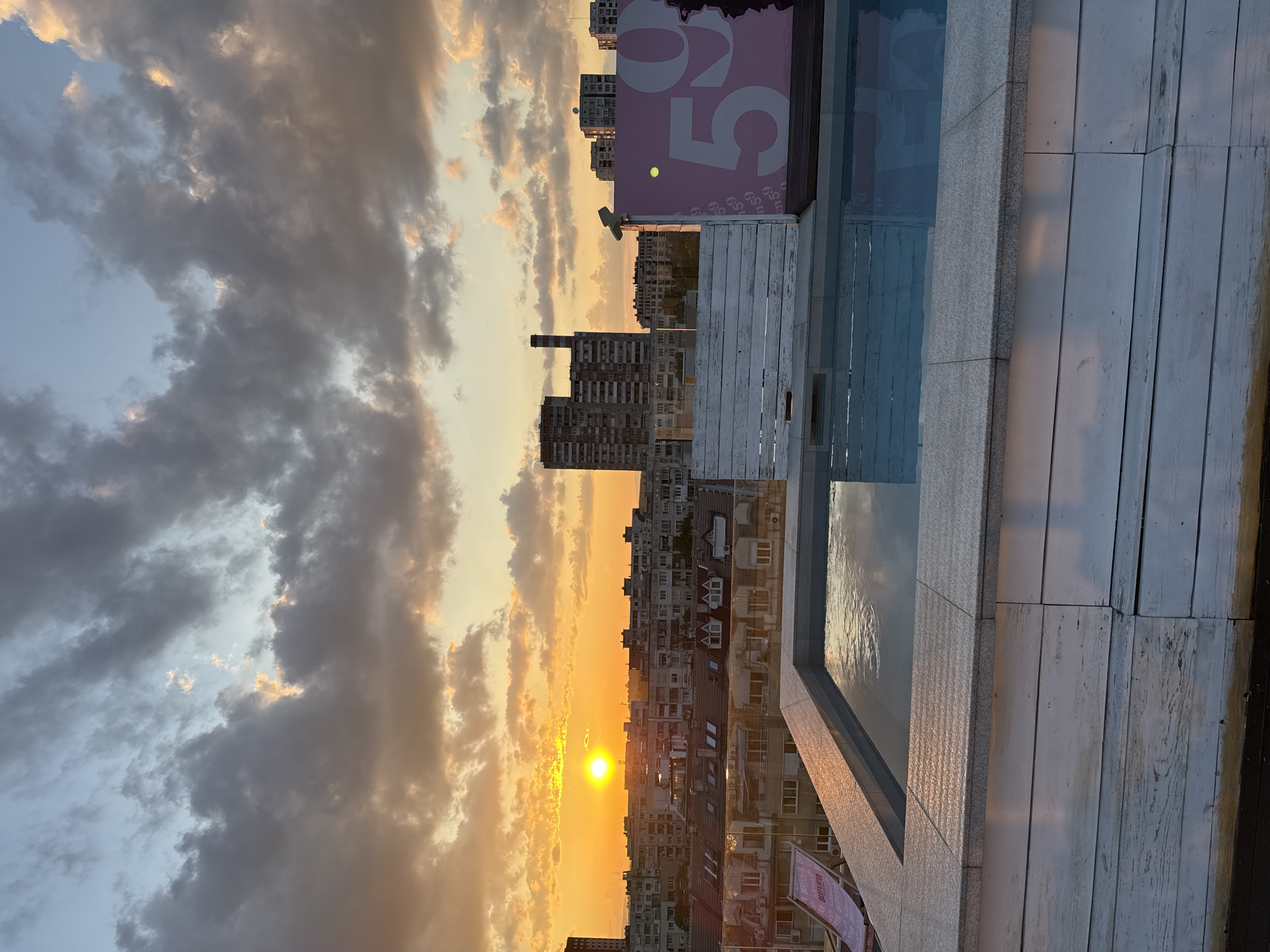 The image captures a rooftop view at sunset in Sofia, Bulgaria. In the foreground, a rectangular plunge pool with a white wooden paneled base and grey granite coping reflects the sky. To the left, a partially visible pink foldable lounge chair bears "BEEFEATER" branding. A white wooden partition or fence separates the pool area. Beyond the rooftop, a dense urban landscape features numerous multi-story residential buildings, ranging from older panel blocks to structures with pitched roofs. A prominent, tall, slender industrial chimney or ventilation shaft is visible among the background buildings. On the right, a large pink sign with white numerals, distinctly showing "59," is partially obscured by the wooden partition. The sun, a bright orange disk, is low on the western horizon, casting a warm, golden illumination across the scene. The sky is filled with clouds, exhibiting colors from dark grey to vibrant orange and yellow where illuminated by the setting sun, with patches of blue sky visible above. No people are present in the scene.