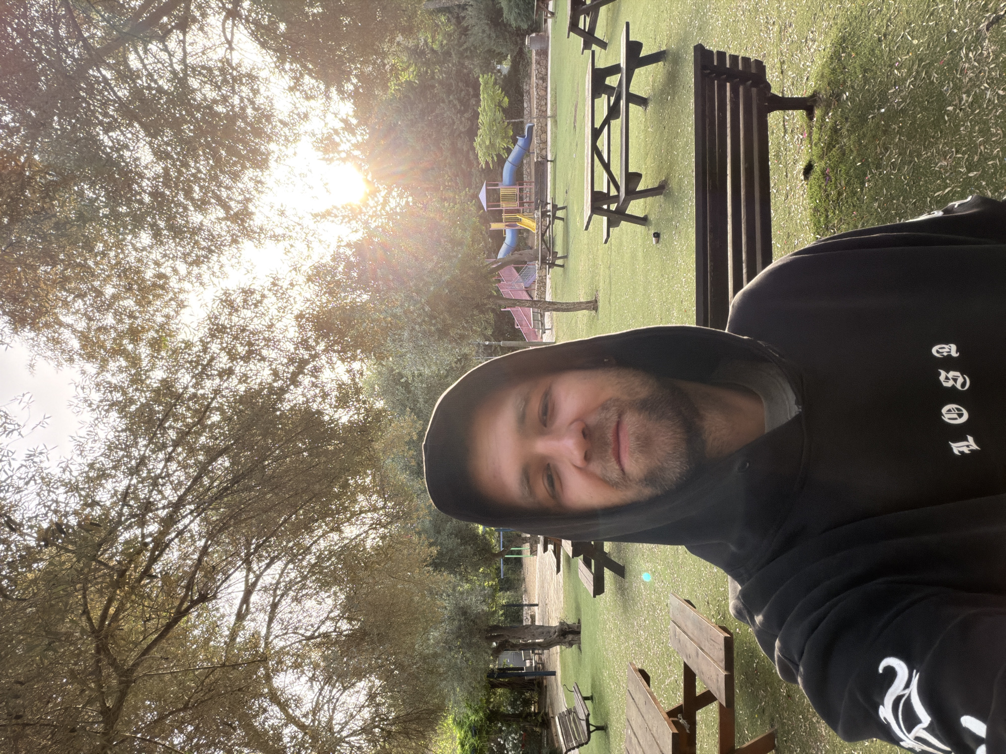 A man wearing a black hoodie is the primary subject, with the background depicting a park setting. The location includes trees, picnic tables and benches, a playground with slides, and artificial grass. The sun is visible through the trees, creating a bright, backlit effect. The man is positioned towards the right side of the frame, with the park scene extending behind and to the left of him. The image is rotated 90 degrees clockwise. The location in the city is unknown.