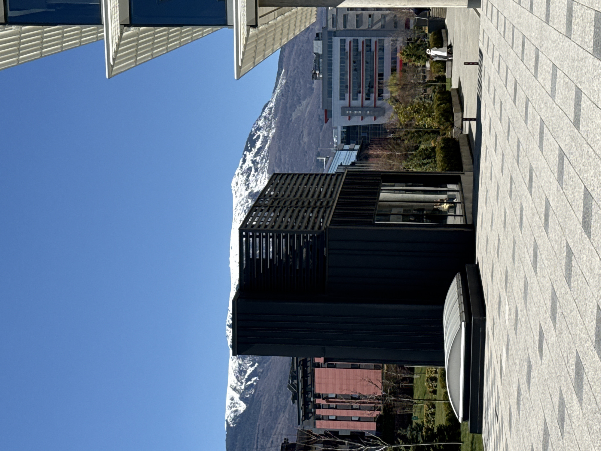 The image shows a modern cityscape under a clear blue sky. The foreground is dominated by a building with dark, grid-like architectural features, next to another building with red brick walls and windows. A tiled pathway with a distinct geometric pattern is visible on the right. In the background, snow-capped mountains loom over the city, a tall building with a white facade and red accents rises. A figure in white stands along the pathway near the building. The scene is located in Sofia, Bulgaria, as indicated by the buildings and mountainous backdrop.