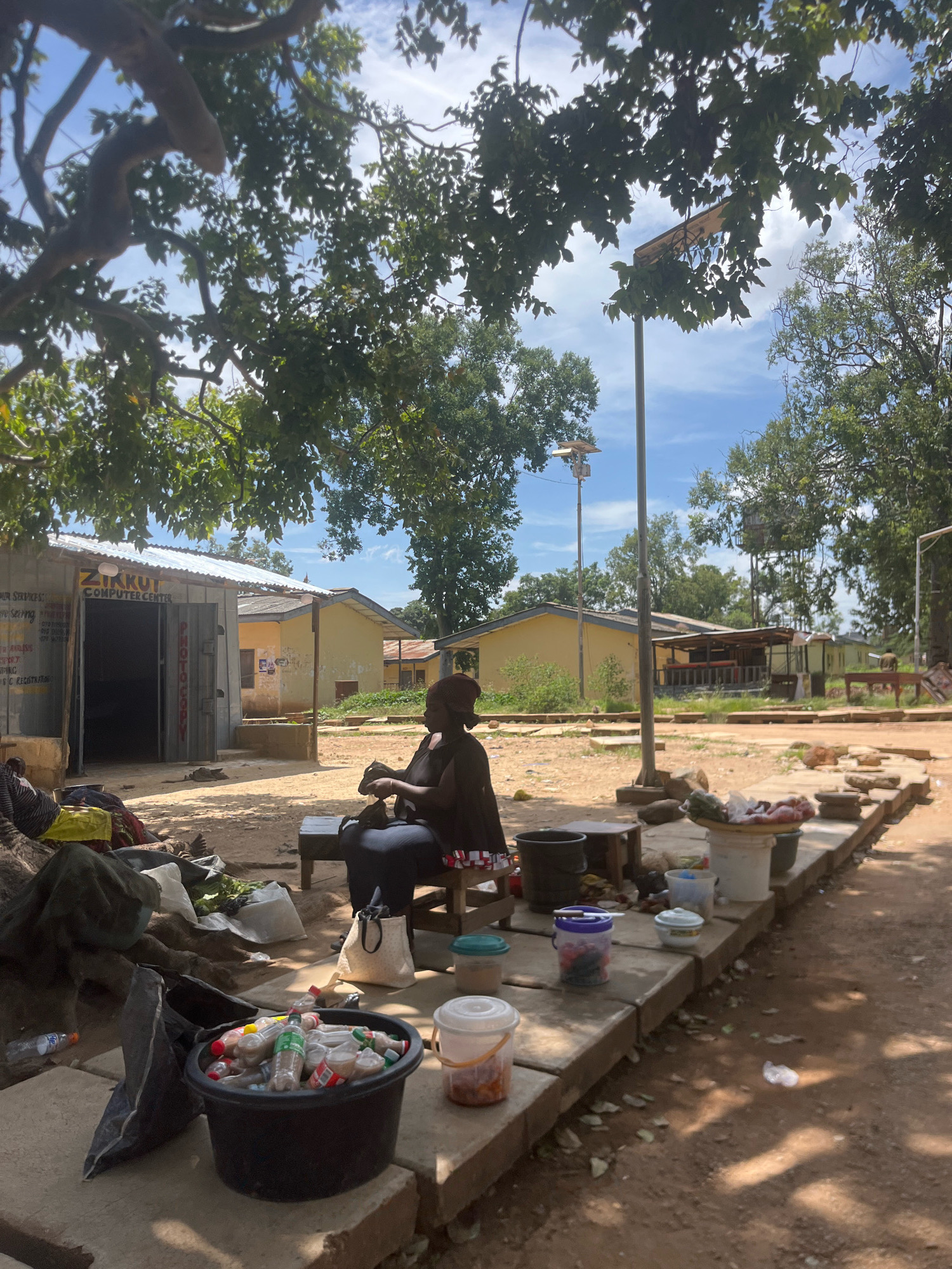 The image captures an outdoor scene in the city of Rafin Par, Nigeria. In the foreground, a woman is seated on a stool on a raised concrete platform, with her back partially to the camera. She is wearing a dark top and a headscarf and appears to be engaged in an activity with her hands, possibly preparing something. In front of her, on the platform, are several containers, including a white bucket filled with produce such as tomatoes and peppers. To the left of the woman, a large black bucket is filled with plastic bottles, many of them containing liquids. Scattered around the base of the platform are other items, including bags and debris.
Behind the woman and the platform, a corrugated metal building is visible, with a sign reading "ZIKKUT COMPUTER CENTER" and "PHOTO COPY". The entrance to the building is open, revealing a dark interior. Further in the background, there are other low-rise buildings with yellow walls and brown roofs, suggesting a residential or commercial area. The scene is shaded by large trees with green foliage, and the ground is a mixture of dirt and scattered leaves. The sky is visible through the trees, with some clouds present. The overall impression is a bustling, informal market or service area.