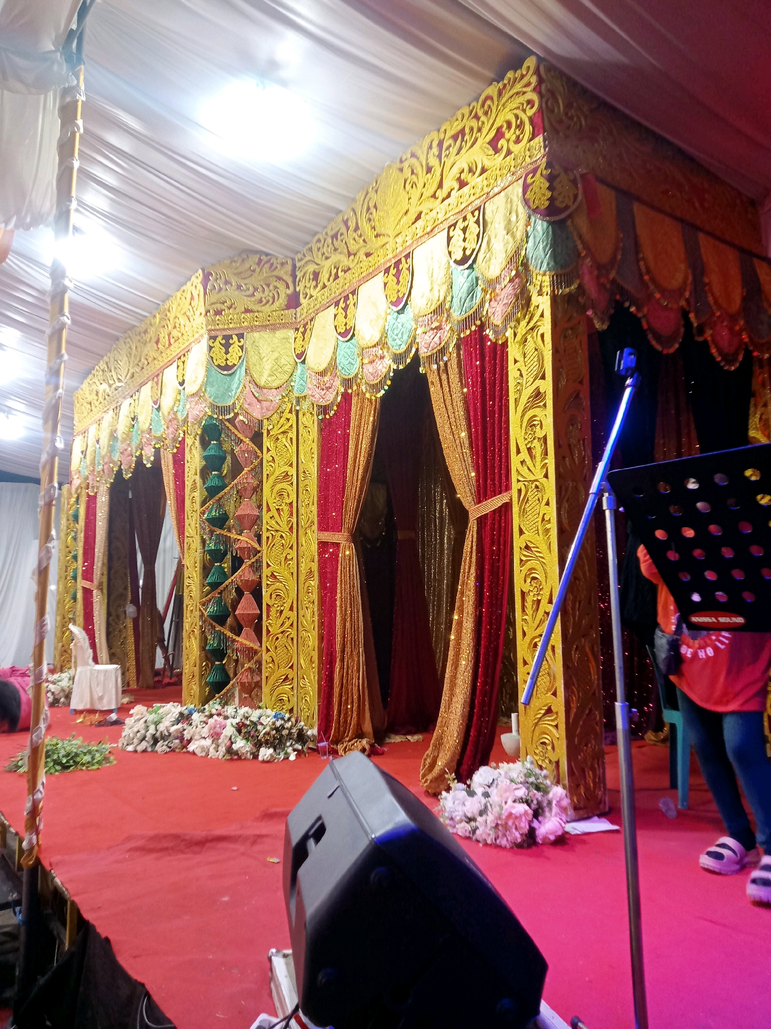 The image depicts a wedding reception setup, likely in Mongolato, Indonesia. The foreground shows a red carpet covering a stage or elevated platform, with a black speaker on the right. A microphone stand with a blue light is also visible, supporting a black music stand. Behind the stand, a person wearing an orange shirt and dark pants is partially visible. The background features an elaborately decorated wedding canopy, adorned with gold filigree, red and gold curtains, and colorful fabric drapes. Floral arrangements are placed on the carpet near the canopy's entrance. The overall scene suggests an indoor event with tent-like coverings and artificial lighting.