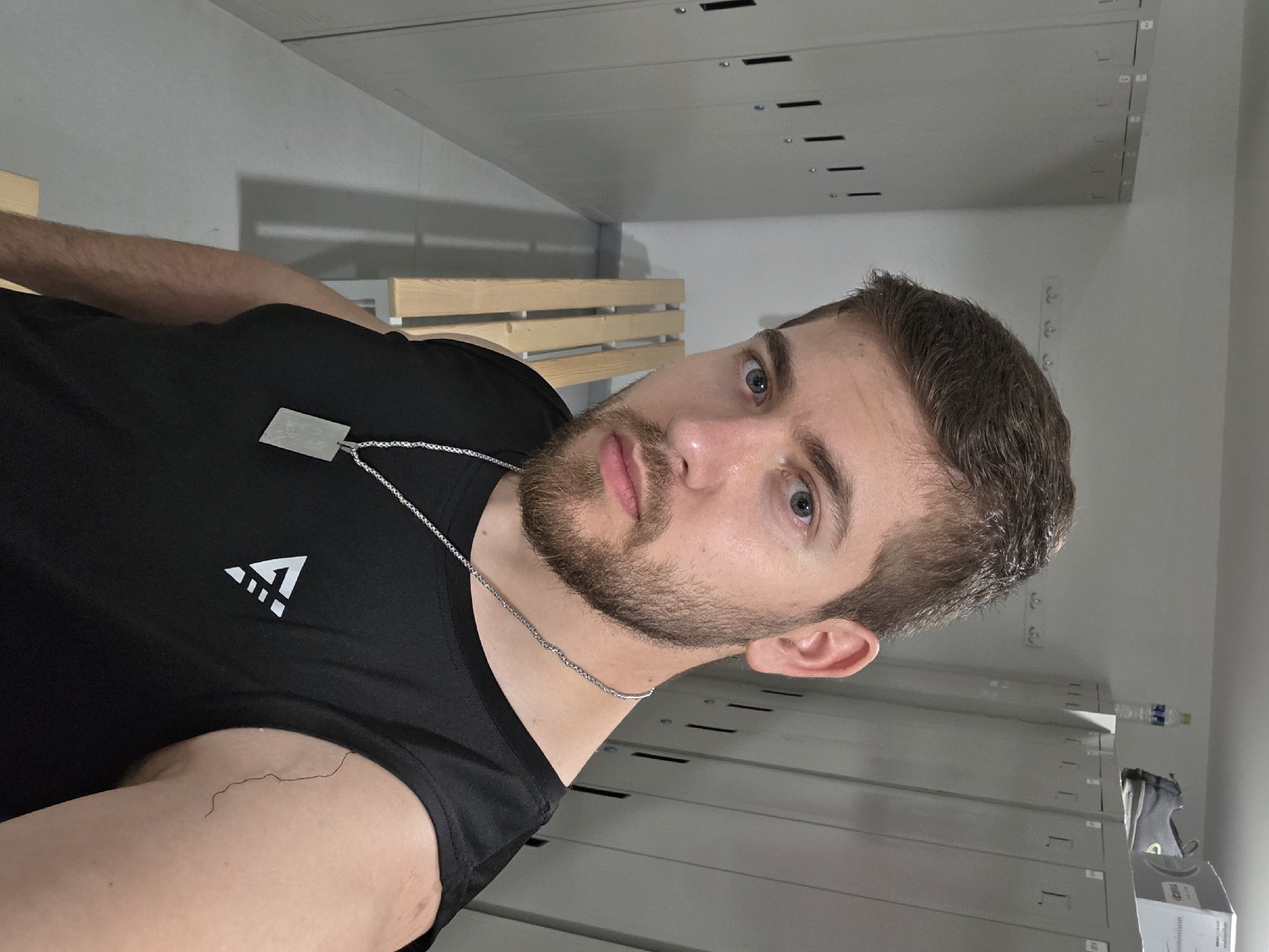 A man stands in what appears to be a locker room. The walls are a light gray color. There are lockers behind him and a wooden bench to his side. The man is wearing a black t-shirt with a white logo and a silver necklace with a pendant. He has a beard and short brown hair. The specific location in Plovdiv, Bulgaria, cannot be determined from the image.