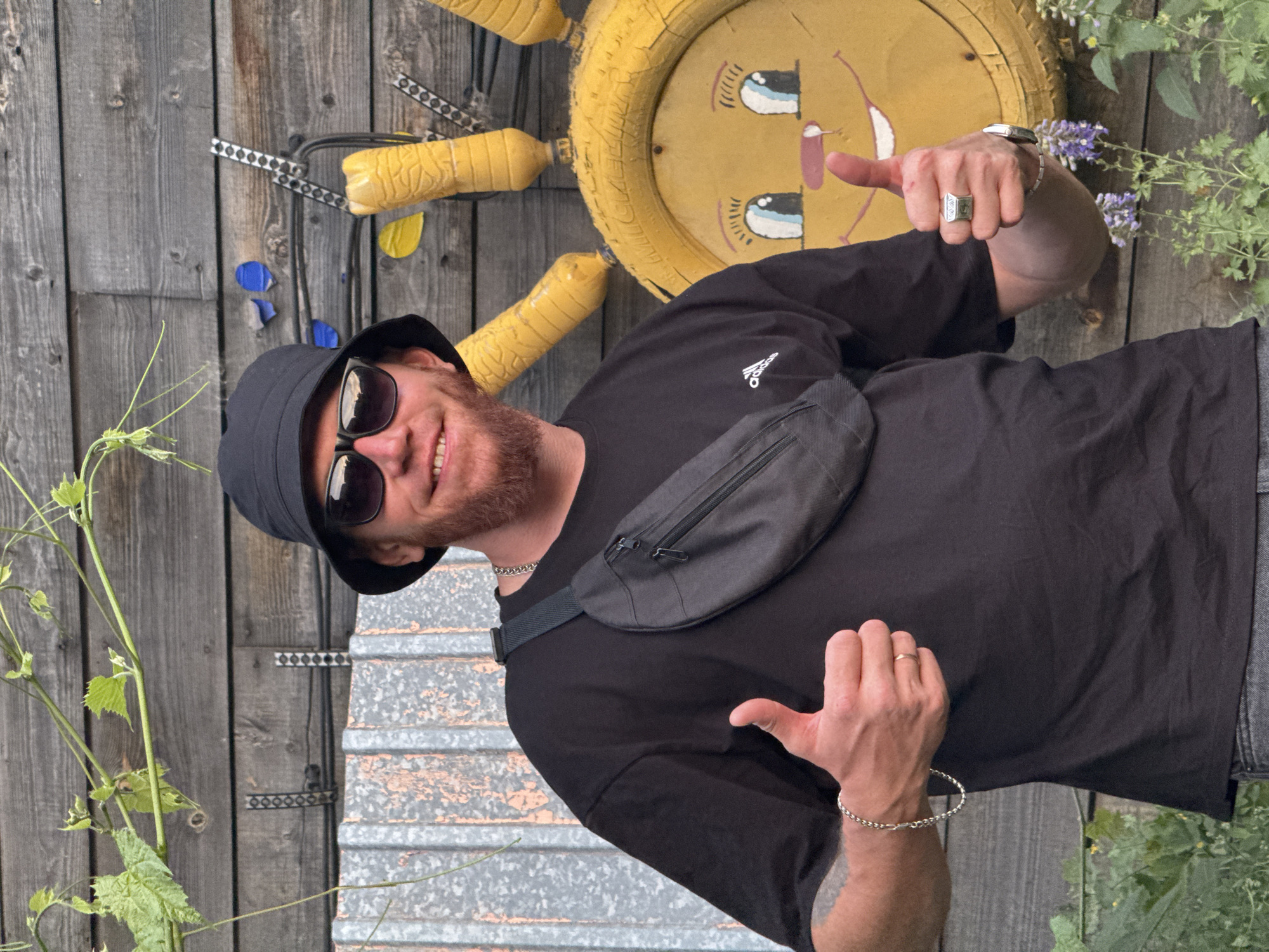 A man wearing a black bucket hat, sunglasses, and a black t-shirt with a fanny pack across his chest is standing in front of a wooden wall. He has a reddish beard and is smiling. He is giving two thumbs up. To his right, there is a yellow, sun-like decoration made from recycled materials, with painted eyes and a mouth. The background is a rustic wooden fence with some greenery and vines growing on it. The man is wearing a silver bracelet and a ring on his right hand. The scene appears to be outdoors in what could be a backyard or a public space with artistic decorations. The specific location in the city of Pavlodar, Kazakhstan is not discernible from the image itself.