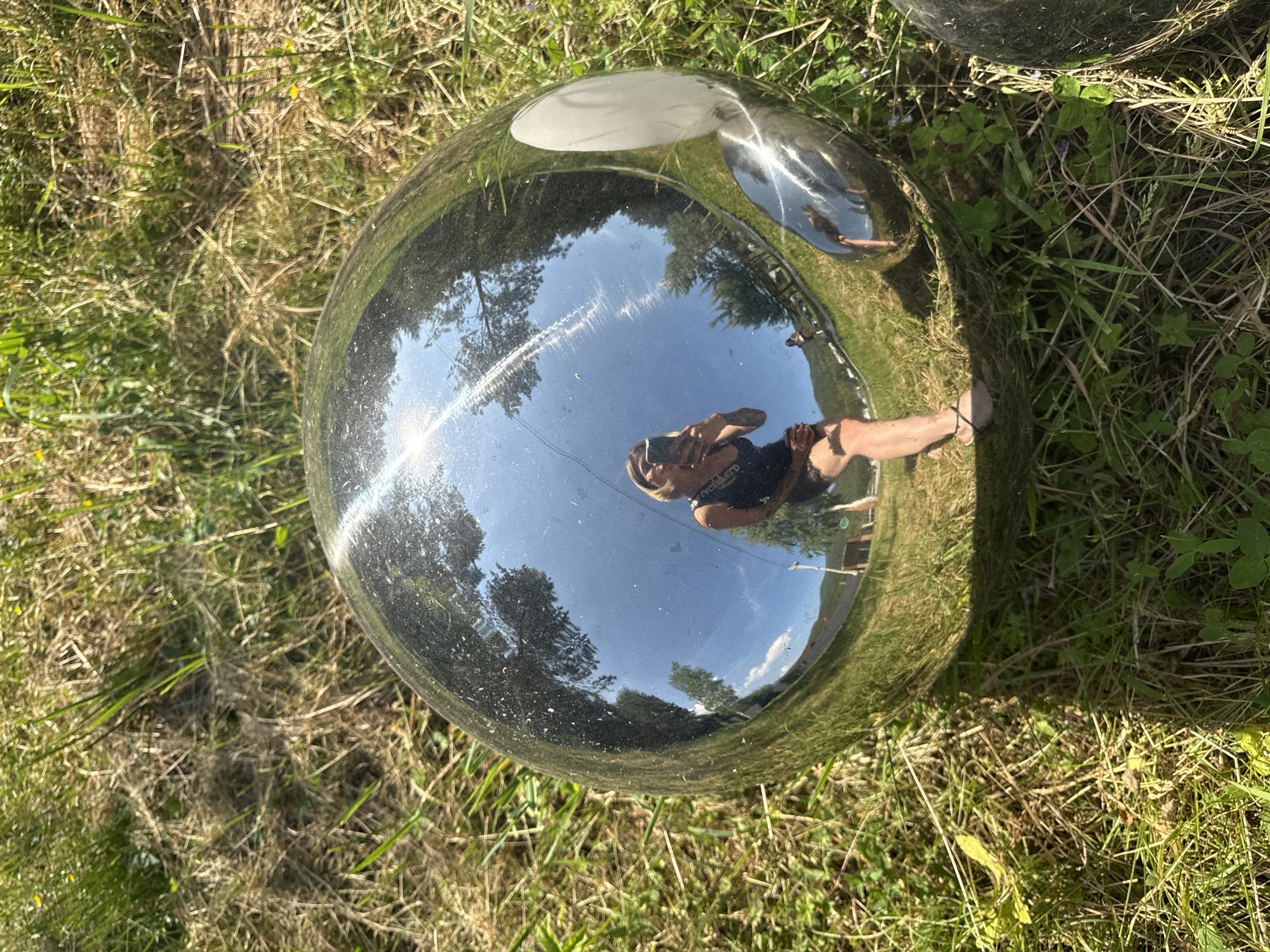 This image depicts a reflective sphere sitting on the ground amidst dry and green grass under bright sunlight. The sphere reflects a scene of a woman holding a phone to take a picture, with a background of trees and a clear blue sky with some clouds. Further in the reflection, a person can be seen walking in the distance, and a hill or mountain range is visible behind them. The overall environment appears to be outdoors, possibly in a park or rural area. The location provided, Ust'-Muny, Russia, is a village.
