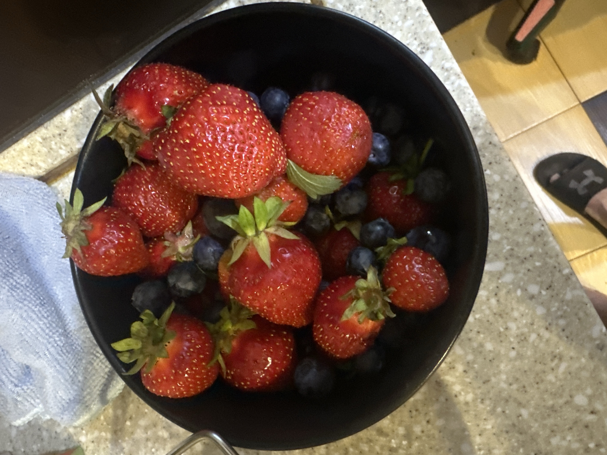 An overhead shot shows a black bowl overflowing with strawberries and blueberries. The bowl sits on a speckled countertop, and a blue patterned cloth is visible in the upper left corner. The lower right of the image shows a foot wearing a black sandal with an "H" logo, and a portion of a dark surface. The scene is indoors, likely a kitchen, and there is no indication of the specific location within Kyiv, Ukraine.