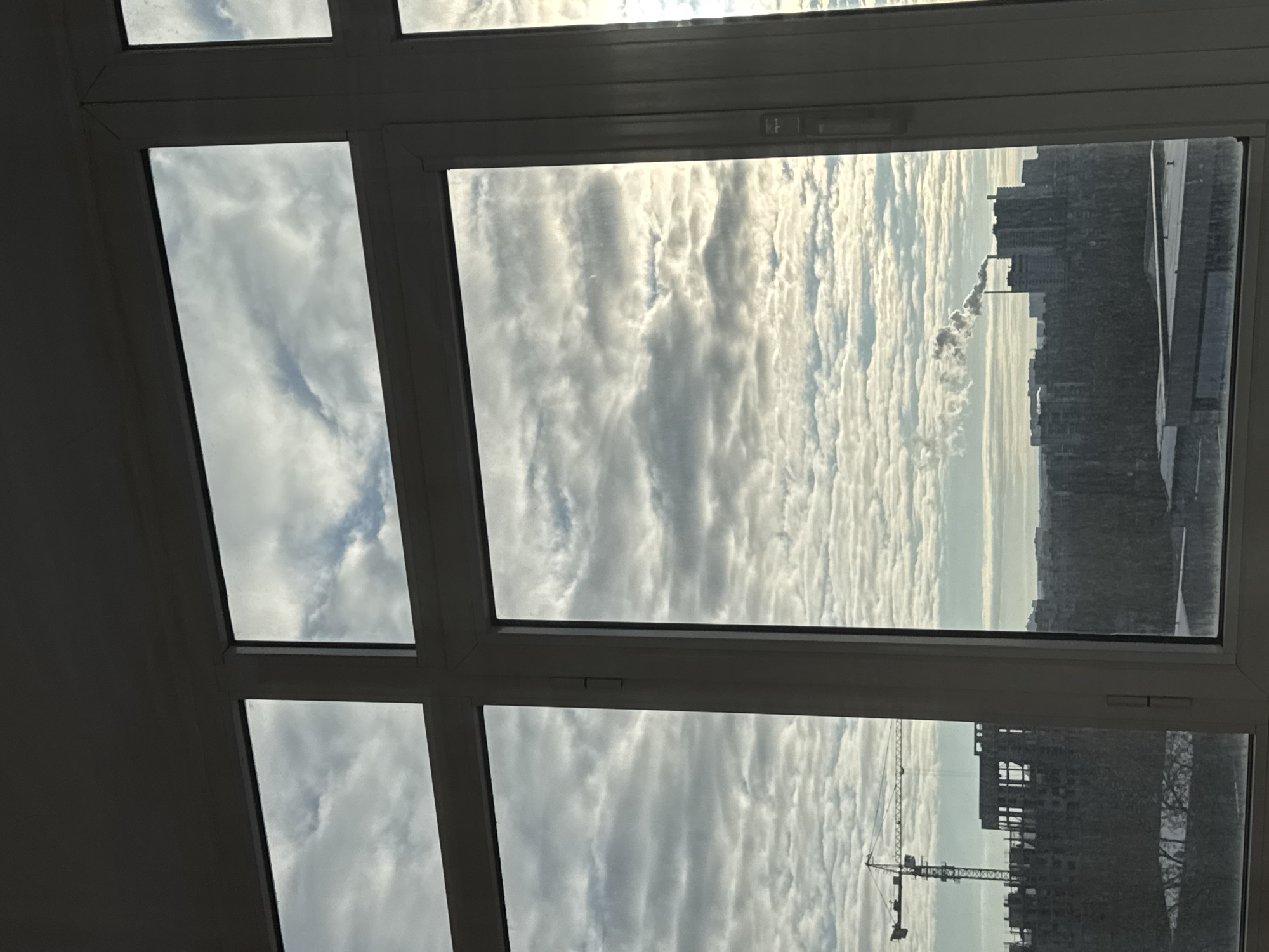The image depicts a view from a window, likely in an apartment or office building. The window frame is white and divides the view into multiple panes. Through the window, the sky is visible, filled with striated clouds. In the central and right-hand panes, urban buildings are seen, including a building under construction with a crane. There is smoke from a chimney. Based on the information, the location is Perm, Russia.