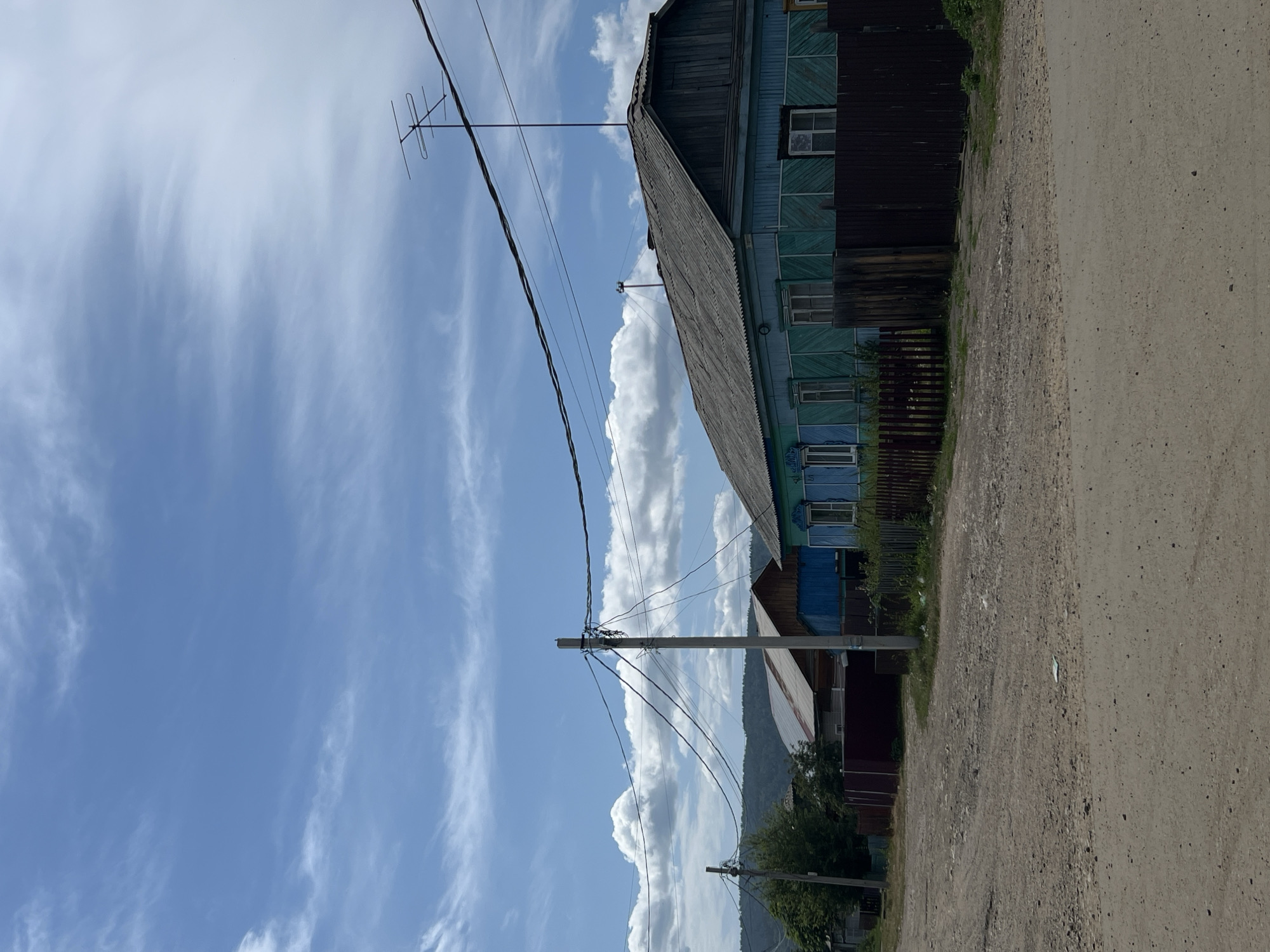 A low-angle, eye-level, outdoor, daytime, full shot depicts a rural dirt road in Bolshaya Rechka, Russia, with several wooden houses and utility poles visible under a partly cloudy blue sky. In the foreground, the dirt road is worn and textured, with scattered small stones and debris. A tall, gray utility pole stands on the right side of the road, adorned with a complex web of black electrical wires that stretch across the frame. To the right of the pole, a long, dark brown wooden fence runs along the property line. Behind the fence, a two-story wooden house painted a vibrant turquoise with green shutters and white window frames is prominent. The house has a weathered, dark gray roof and a wooden attic dormer. An antenna is mounted on the roof. To the left of the turquoise house, a smaller, less distinct wooden house with a silver corrugated roof and a blue facade is partially visible behind a dark red wooden fence. Further left, a dark green tree stands beside another utility pole, also covered in electrical wires. In the background, rolling hills covered in green foliage are visible beneath a sky filled with streaks of white clouds against a bright blue backdrop. The overall scene conveys a sense of rustic village life.