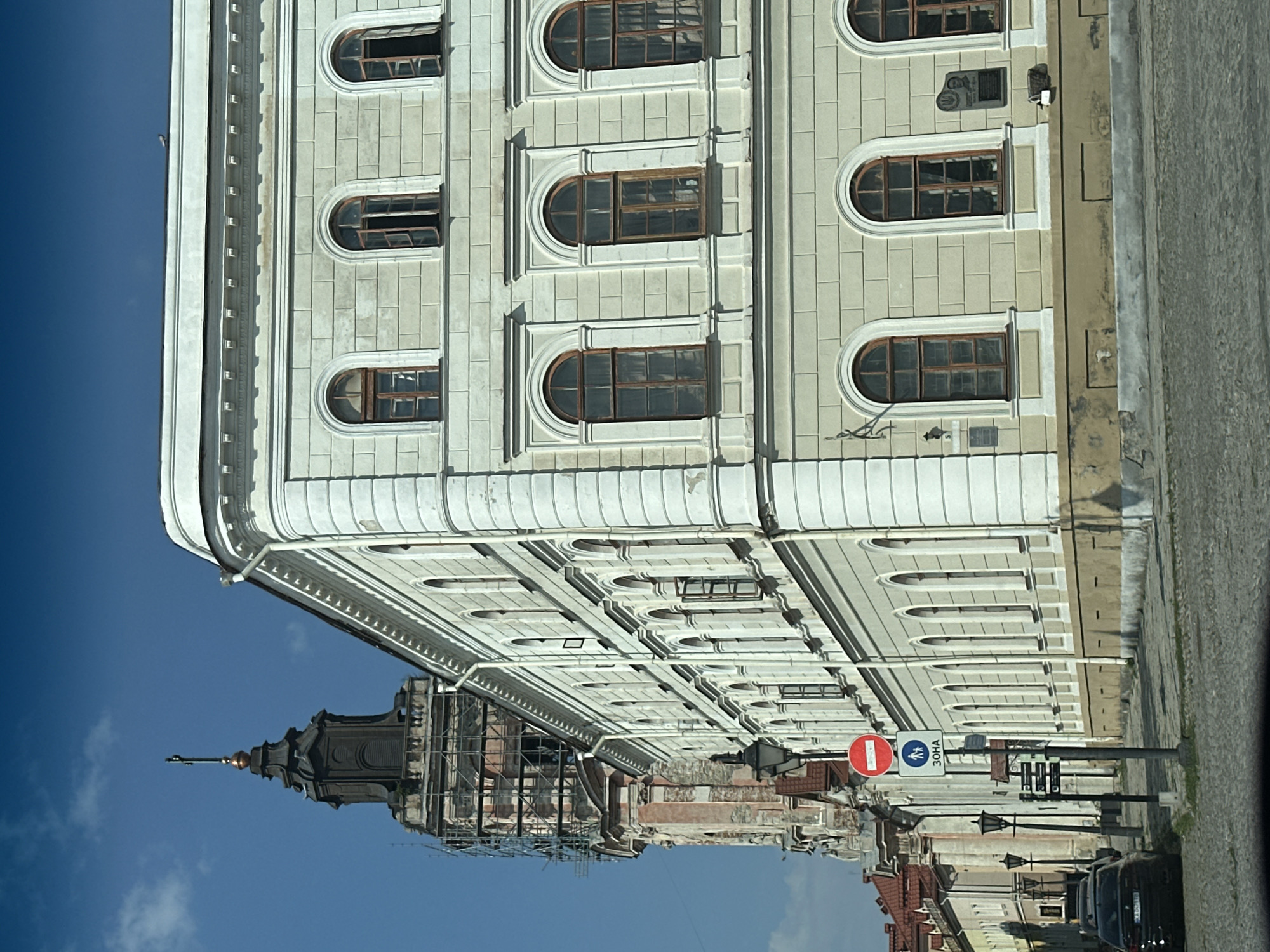 The image shows a view of a light-colored, multi-story building with arched windows on the right side, and a tall, dark tower with scaffolding on the left. The building features classical architectural elements, including columns and decorative moldings. The tower appears to be undergoing renovation. A street with cobblestone paving runs in front of the buildings, with several cars parked along the side. Streetlights and traffic signs are visible along the street. The sky is clear and blue. The overall scene suggests a historical urban environment in Kamianets-Podilskyi, Ukraine.
