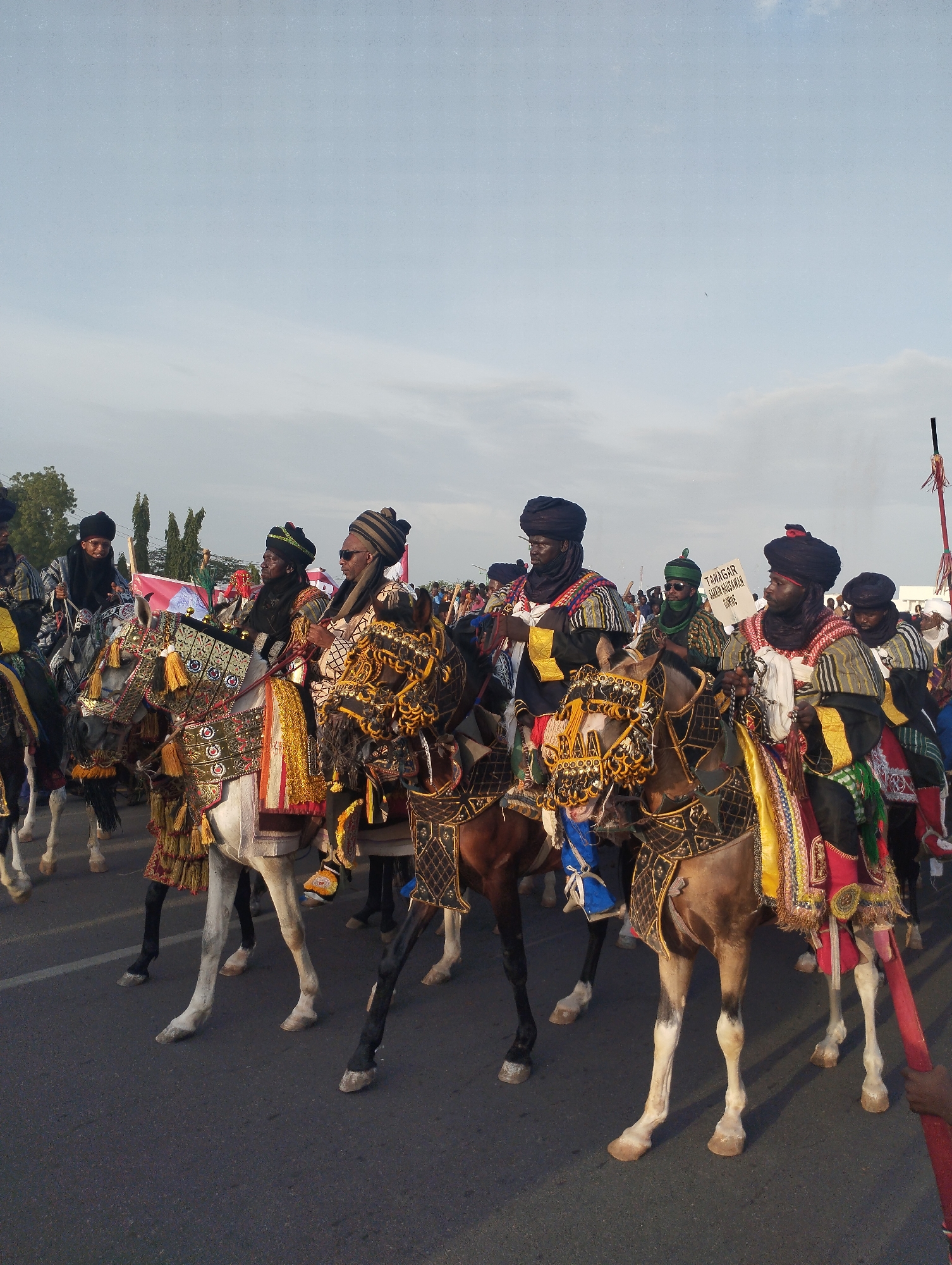 A group of men on horseback are parading down a paved road. The men are adorned in colorful, traditional garb with black head coverings. The horses are elaborately decorated with patterned cloths, tassels, and ornamental headpieces. In the background, other figures and flags can be seen, suggesting a festive or ceremonial occasion. A sign is also visible, partly obscured by the crowd, with the word "TAWAGAR" on it. The scene is set outdoors, likely in the city of Gombe, Nigeria.