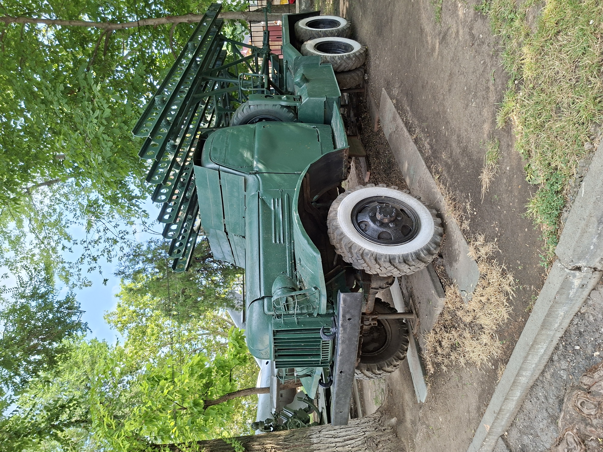 A green military truck with a rocket launcher is parked outdoors on a paved surface. The truck has large tires, a prominent grille, and the rocket launcher is mounted on the back. Trees with green leaves surround the vehicle, and the sky is visible through the branches. In the background, another military vehicle, possibly an airplane, is visible. The scene appears to be a museum or a memorial site.