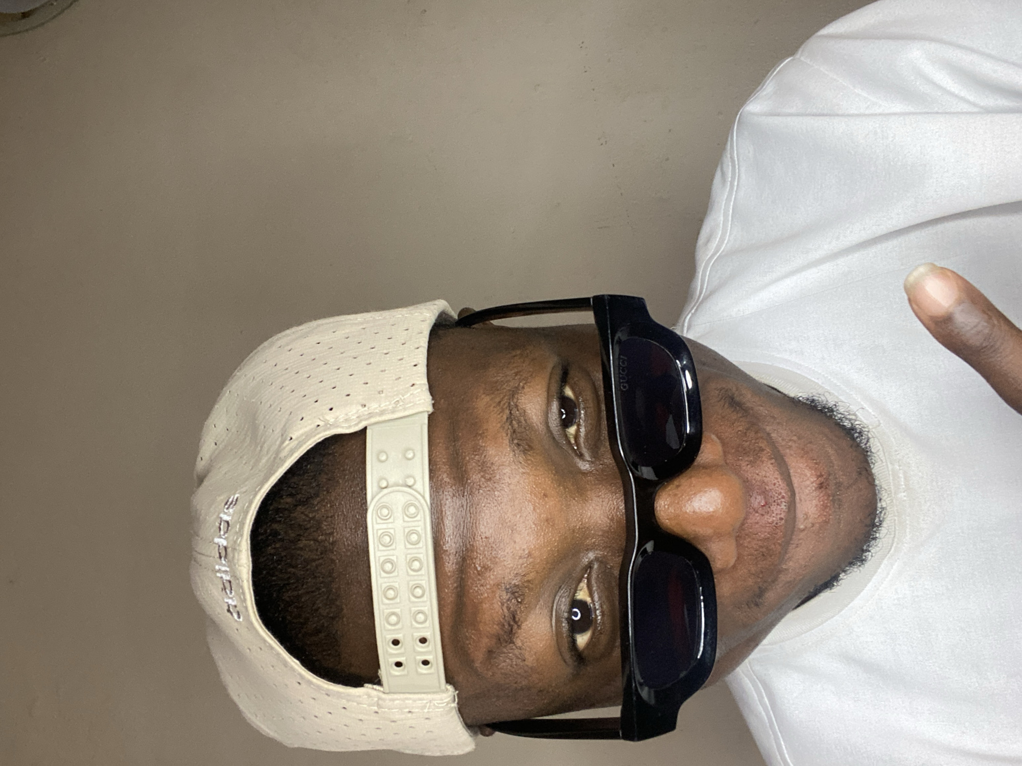 A man is facing the camera. He is wearing a white Adidas hat, black Gucci sunglasses, and a white t-shirt. He is holding up his index finger. The background is a plain beige wall. There is no visible information to indicate that this image was taken in Bukuru, Nigeria.