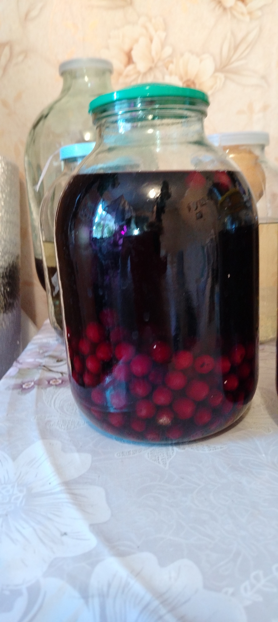 The image shows a large glass jar filled with a dark liquid and numerous red berries, likely cherries or a similar fruit, positioned on a table covered with a light-colored floral-patterned tablecloth. Other glass jars are visible in the background, suggesting a collection or storage of liquids and preserves. The background wall features a floral pattern. The image was taken in Зипуново, Russia.