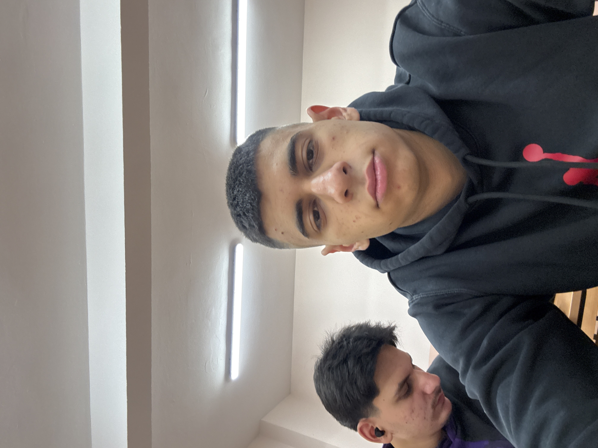 Two young men are in an indoor setting. One, in a black hoodie with a red logo, is closer to the camera. The other is slightly behind and to the left, wearing a purple garment and an earbud in his left ear. They are under bright fluorescent lights, possibly in a school or office environment, judging by the ceiling. The location in Sofia, Bulgaria, cannot be determined from the visual information.